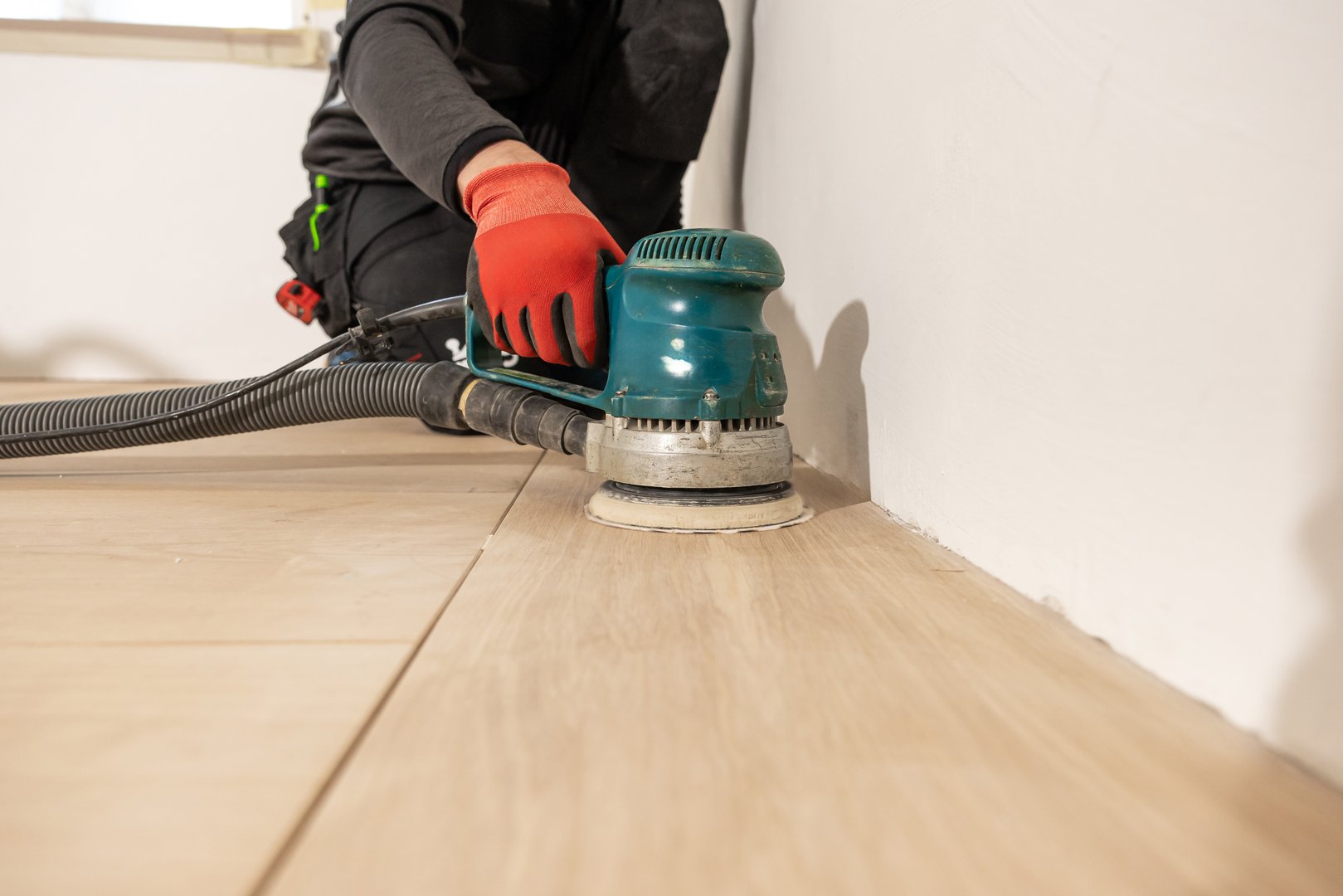 A person in dark work attire uses a green floor sander with a black hose to polish a wooden floor. Natural light enters through a window nearby.