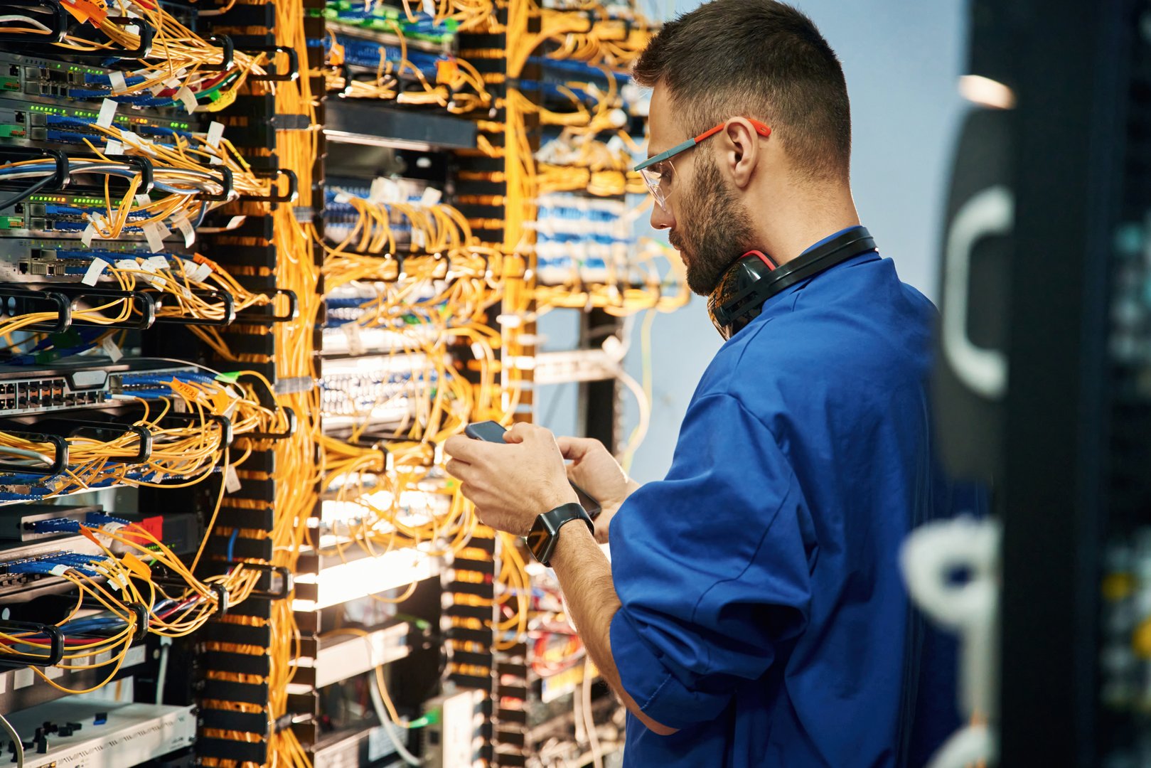 Holding smartphone. Young man is working with internet equipment and wires in server room.