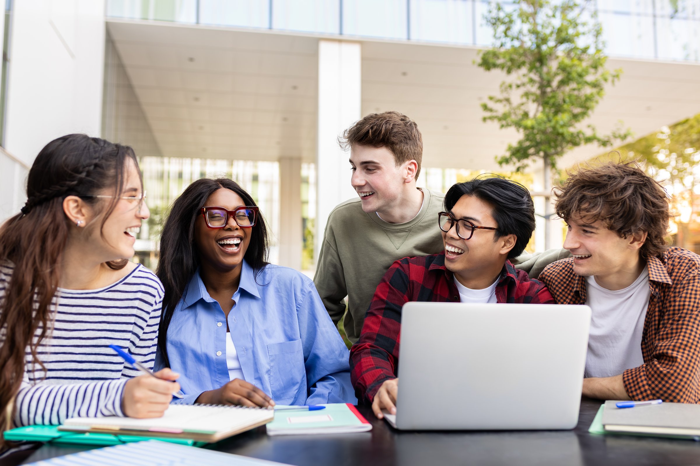 Five cheerful university students are laughing and working together on a project using a laptop outdoors