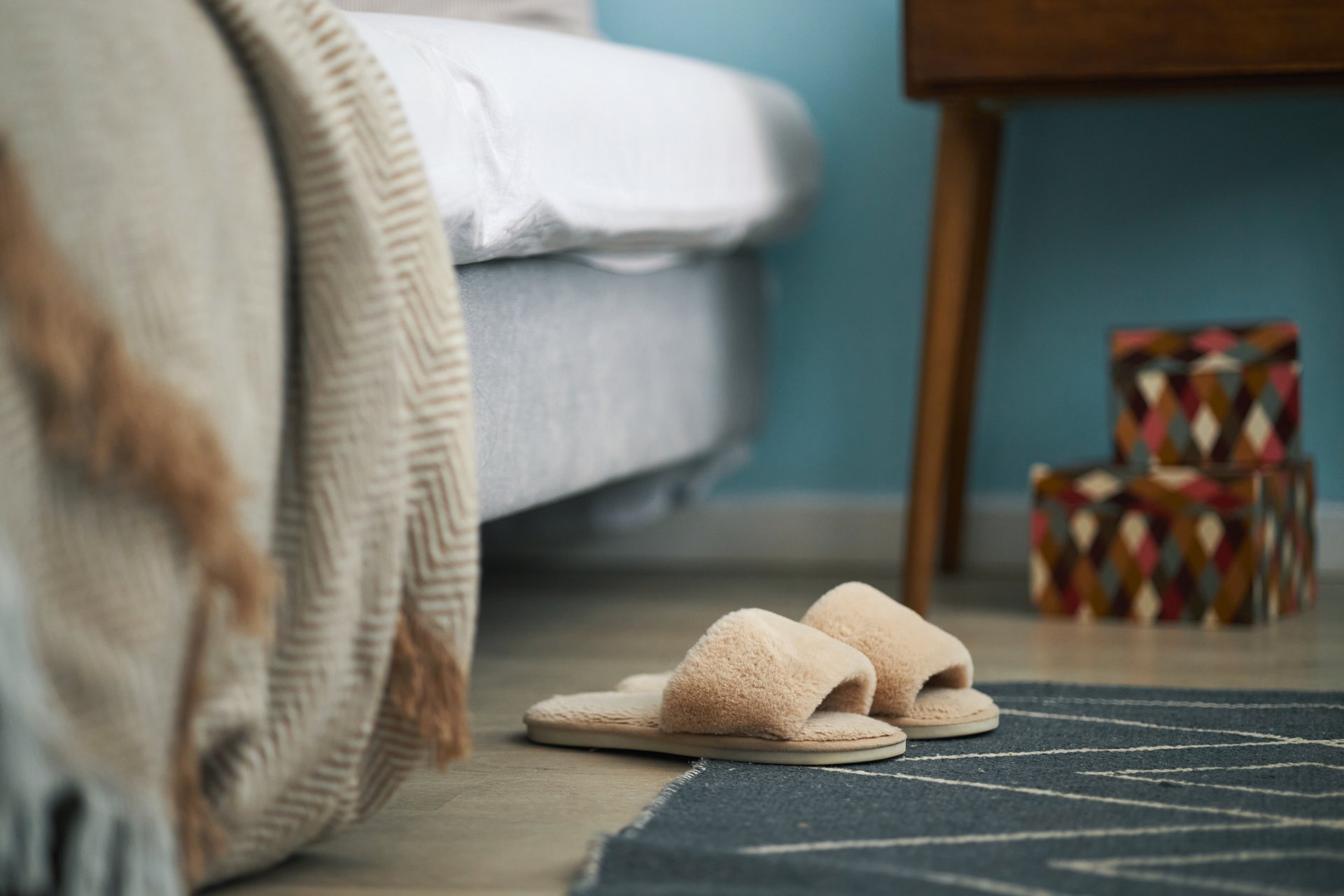 Comfortable-looking bedroom featuring soft slippers placed on rug by bed with a wooden nightstand adorned with colorful boxes visible in background
