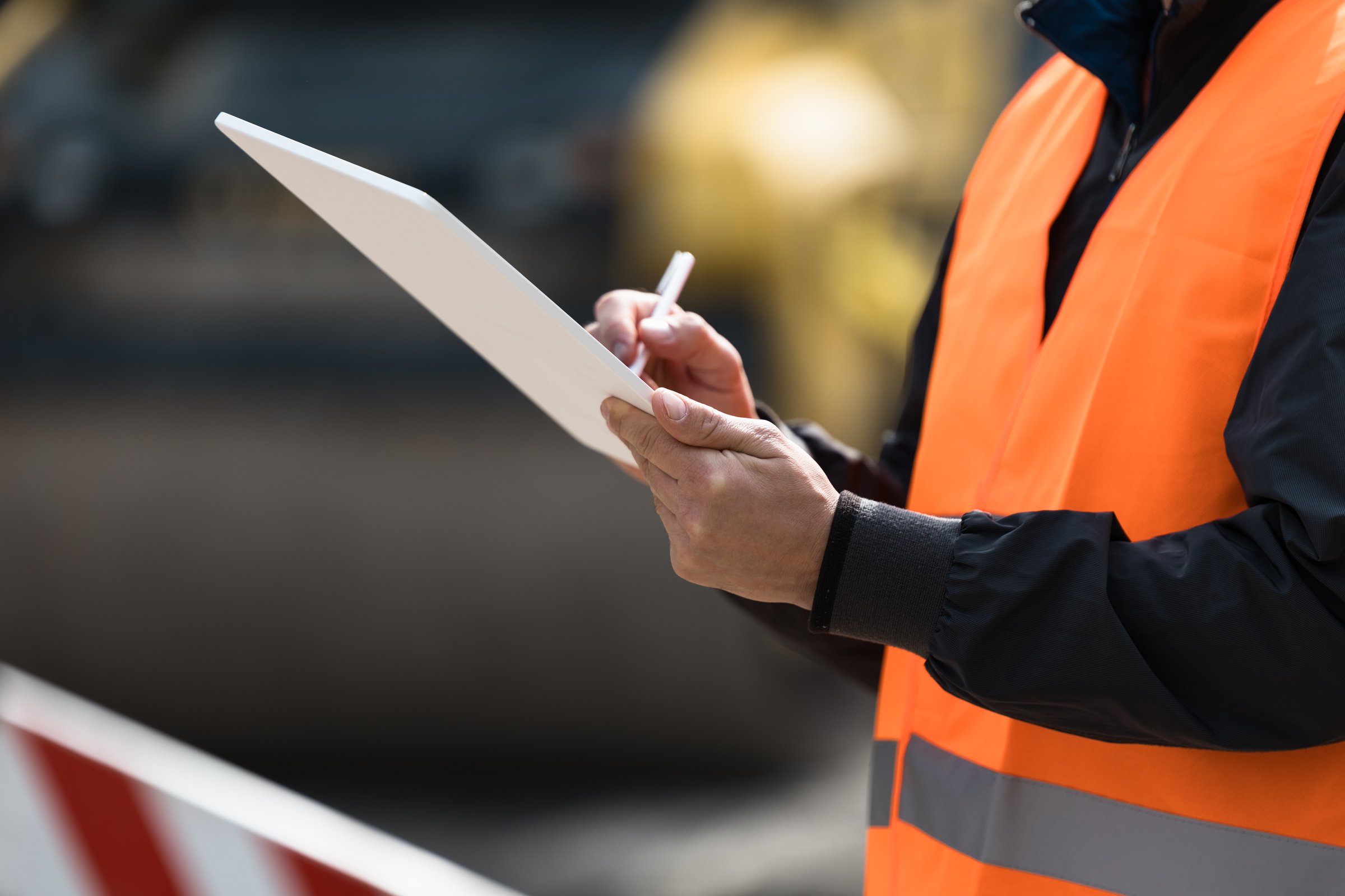 A construction site supervisor in an orange safety vest writes on a notepad, monitoring the work around him amid heavy machinery and ongoing activities.