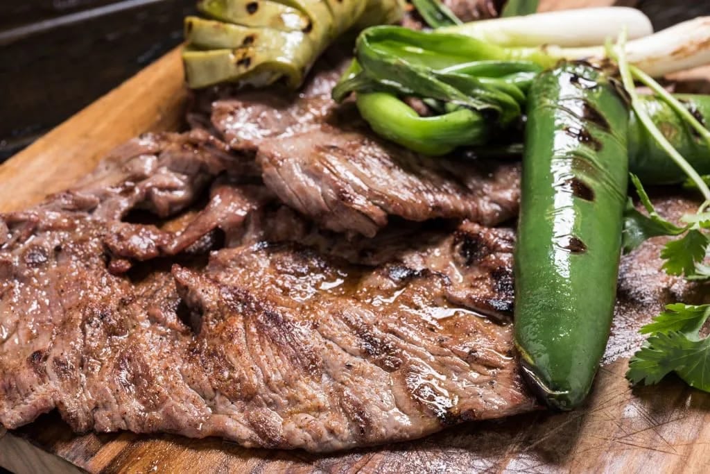 Grilled steak with green peppers and scallions on a wooden board, garnished with fresh herbs.