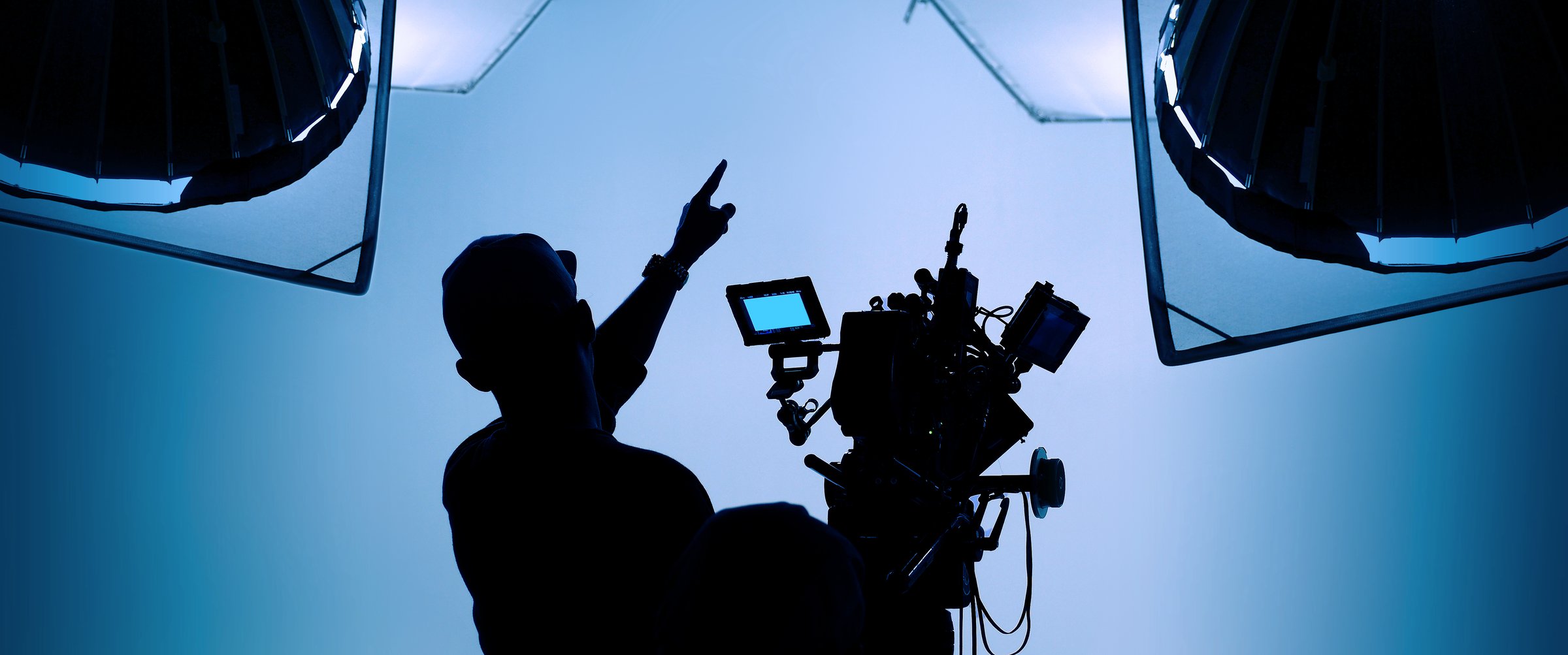 Silhouette of a person pointing near a professional video camera, surrounded by studio lights on a blue background.