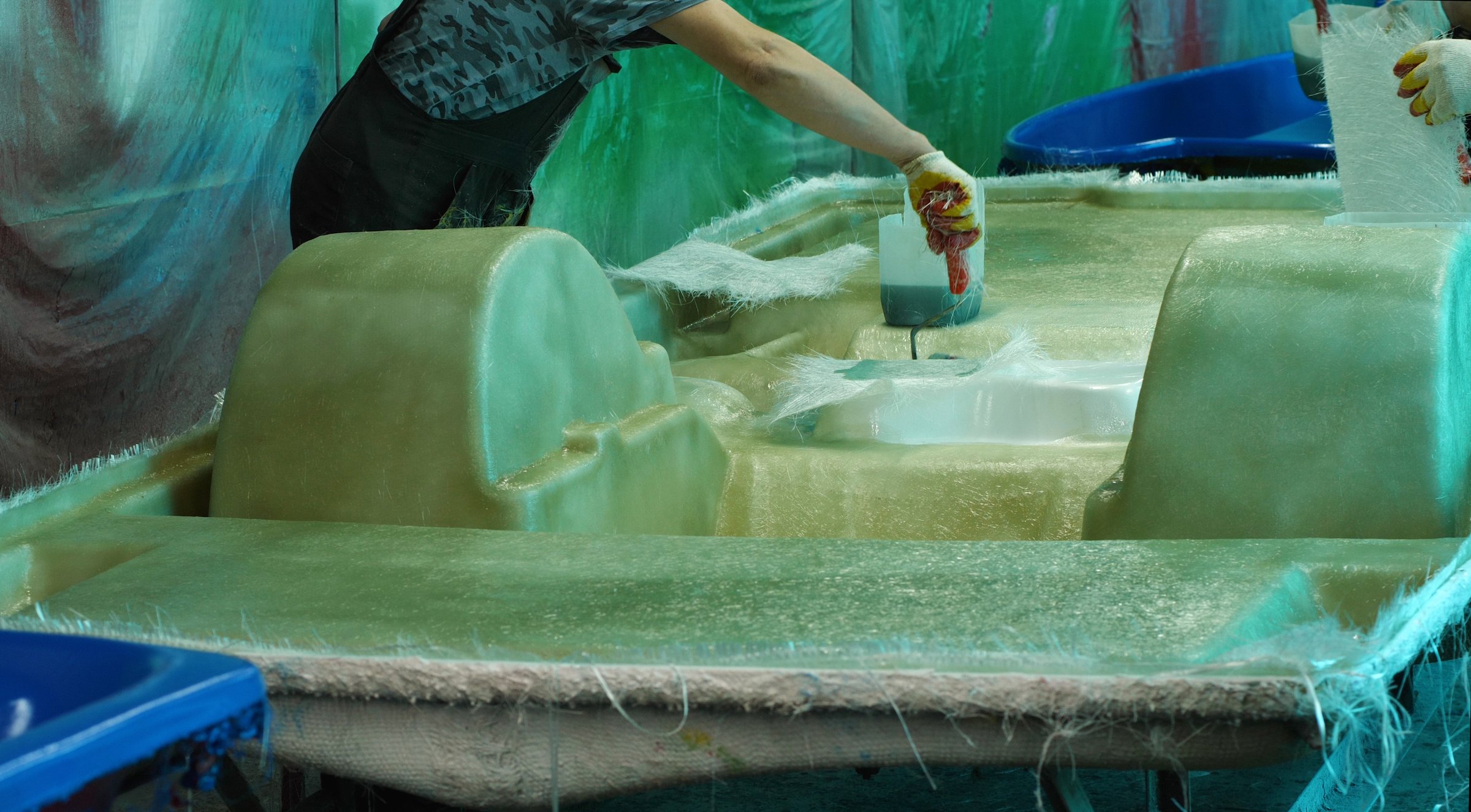 A man uses a paint roller to apply adhesive to fiberglass, gluing the hull of a pedal catamaran. Building boats in a fiberglass shipyard