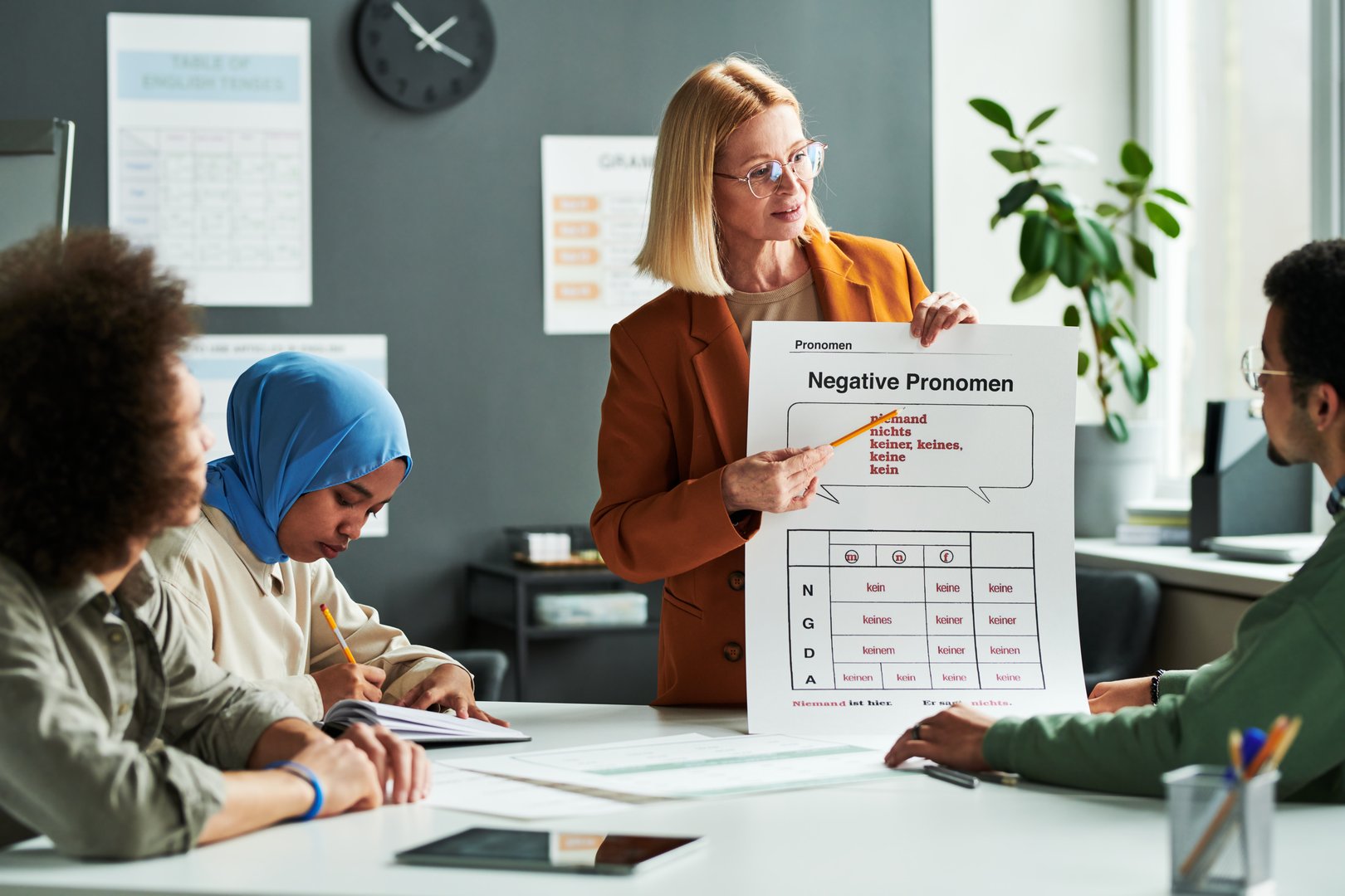 Confident teacher of German language explaining negative pronouns to group of students while pointing at table during presentation