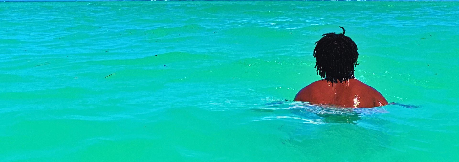Person with dreadlocks swimming in clear turquoise ocean water, viewed from behind.