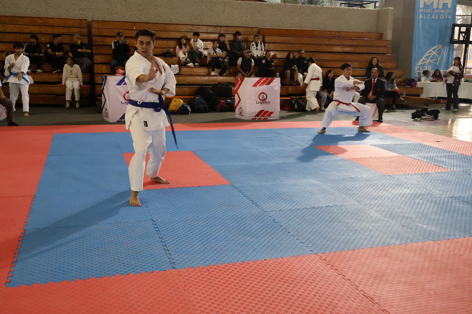 Two martial artists in blue belts perform kata on a padded floor in front of a seated audience in a gymnasium.