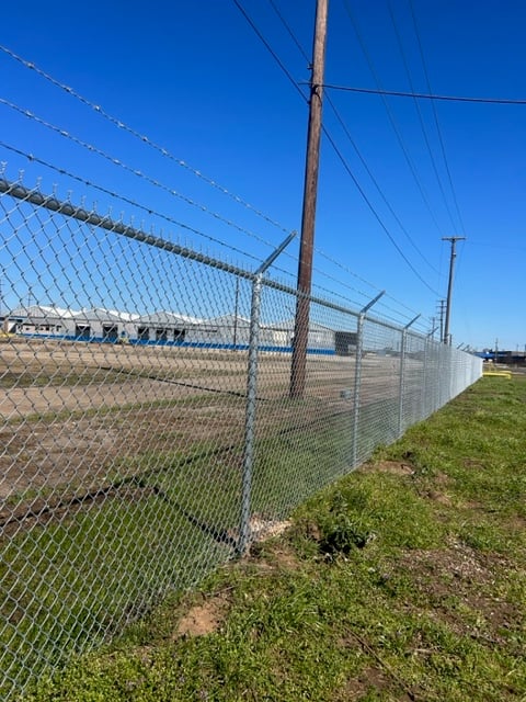 Chain-link fence topped with barbed wire, bordering an industrial area. Clear blue sky and grassy ground in the foreground.