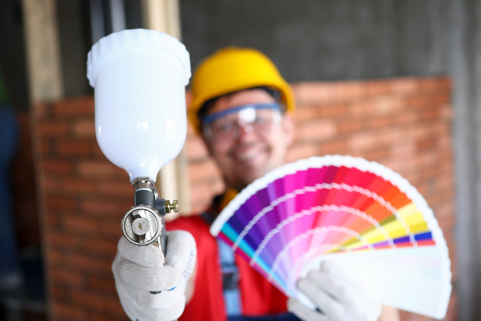 Close-up of smiling construction site worker holding colourful palette with samples and empty professional airbrush gun. Renovation and interior design concept