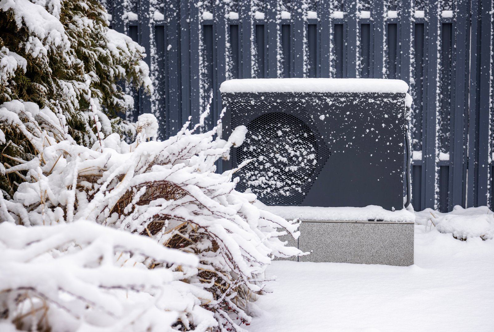 Snow-covered outdoor heat pump unit beside frosted bushes and conifers against a gray metal fence in a winter garden setting. High quality photo
