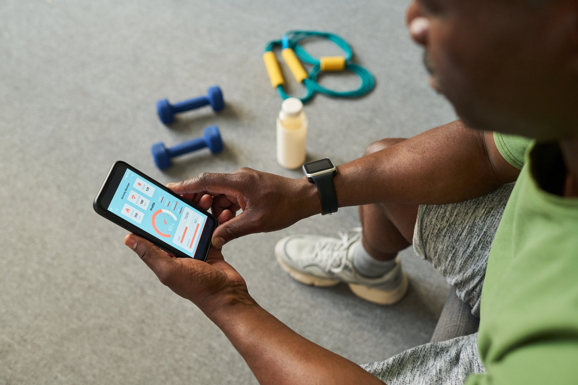 Senior Black man sitting on floor using fitness tracking app on smartphone, holding device with both hands, exercise equipment including dumbbells and resistance band visible nearby