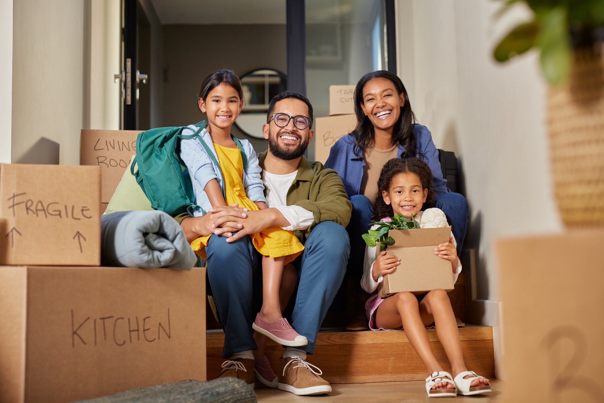 Middle eastern family sitting on stairs surrounded by moving boxes. Indian parents and their two daughters smiling on moving day. Mixed race family with daughters enjoy a moment of relax together during relocation.