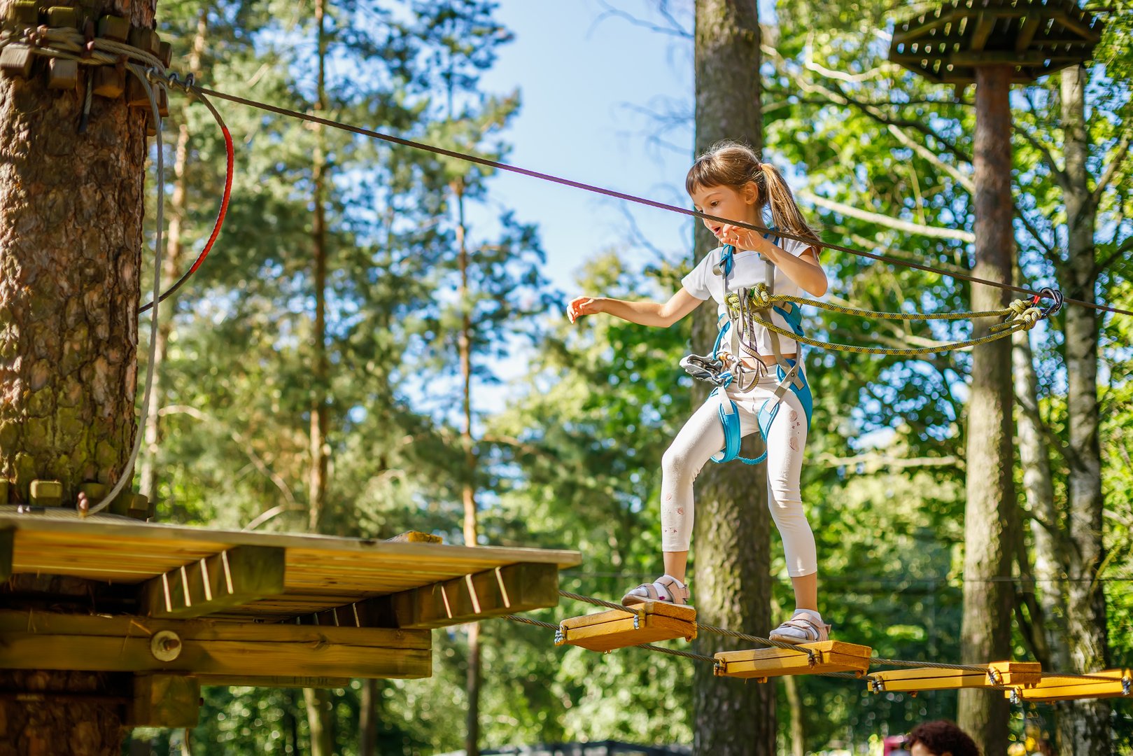 Young girl confidently navigates a challenging rope course high above the ground in a forest setting. Dressed in safety gear, she maintains her balance with focus and determination