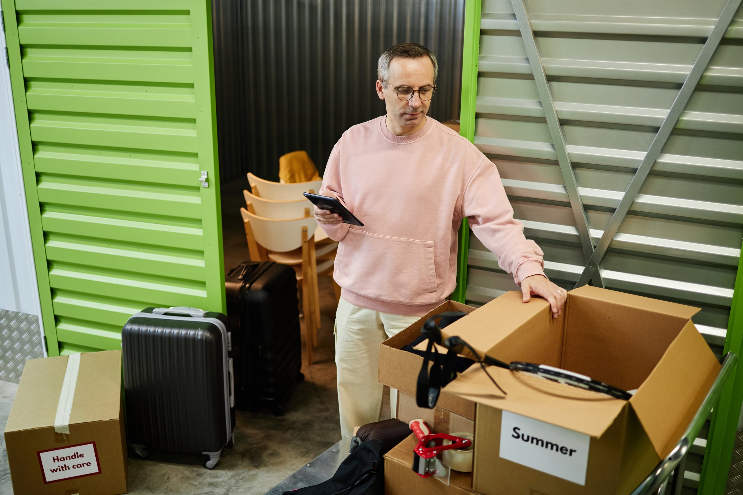 Middle aged Caucasian man organizing cardboard boxes in storage unit while holding tablet, standing near luggage and labeled containers, focused on sorting personal belongings
