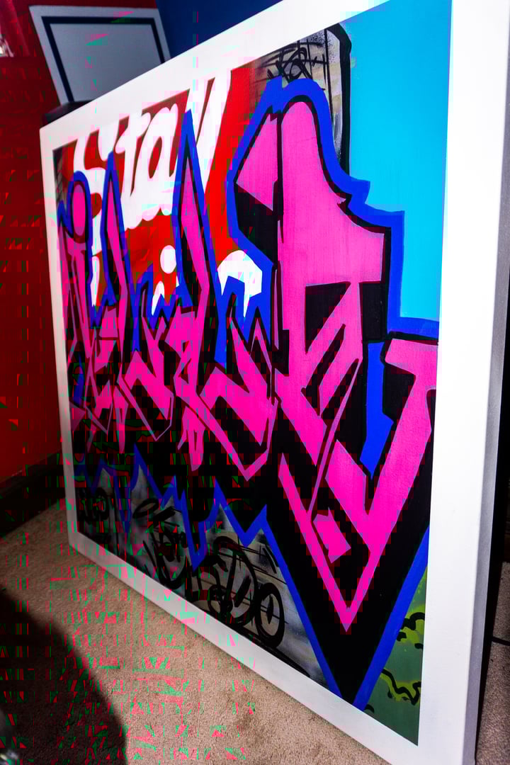 Framed graffiti art with bold pink lettering on a blue and red background, leaning against a wall.