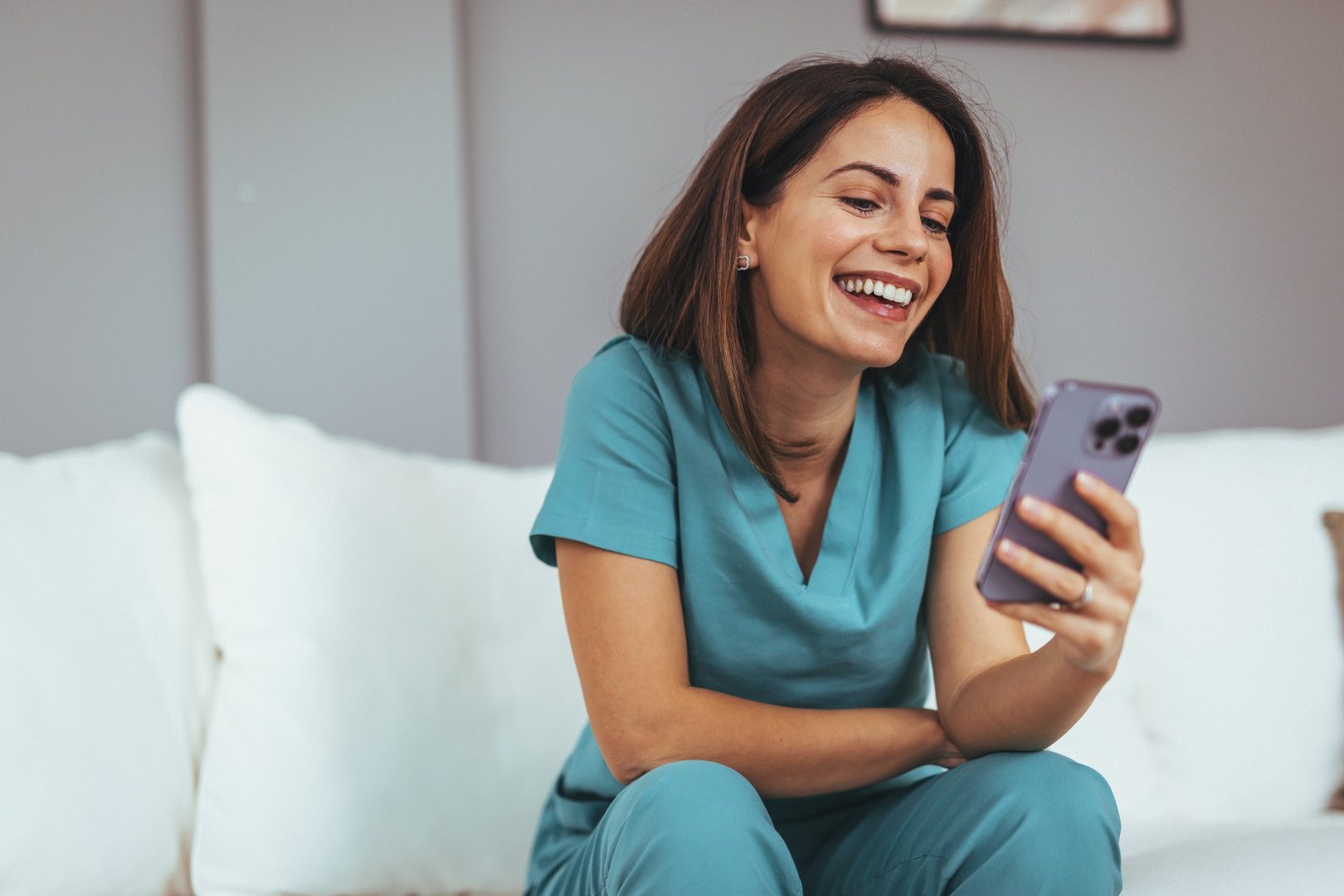 A cheerful woman in medical scrubs happily interacts with her phone, sitting comfortably on a couch in a cozy home setting.