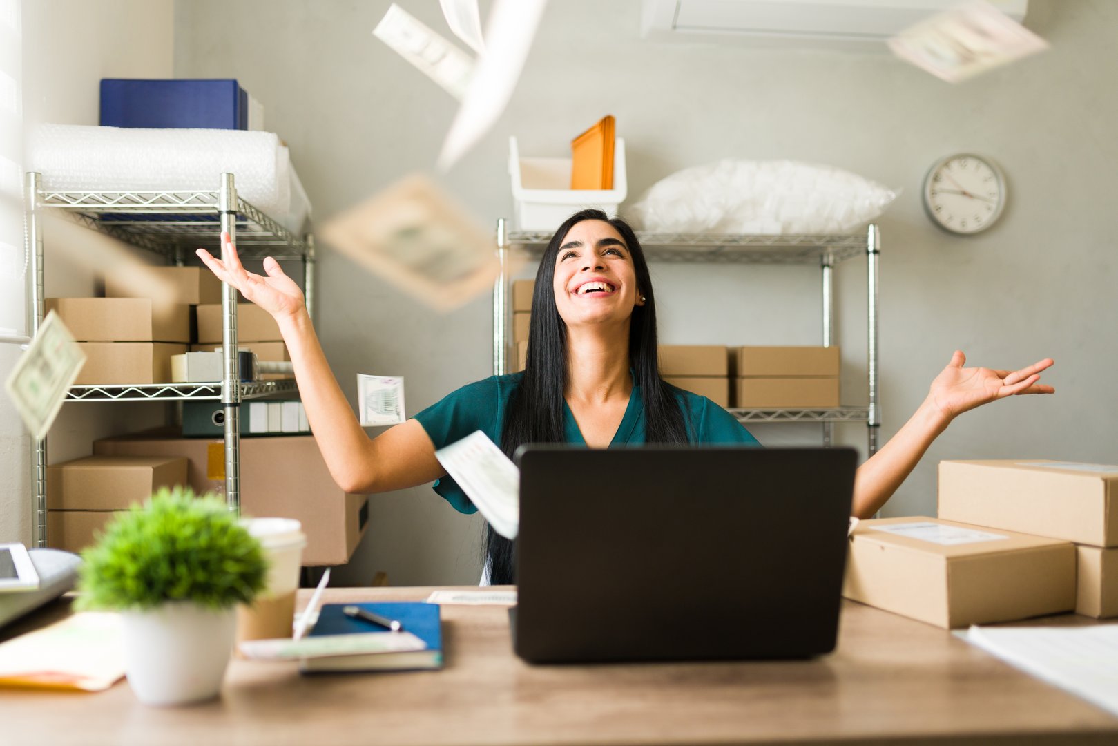 Thrilled businesswoman joyfully tossing money in the air as she works on her laptop in her warehouse, celebrating the success of her small business