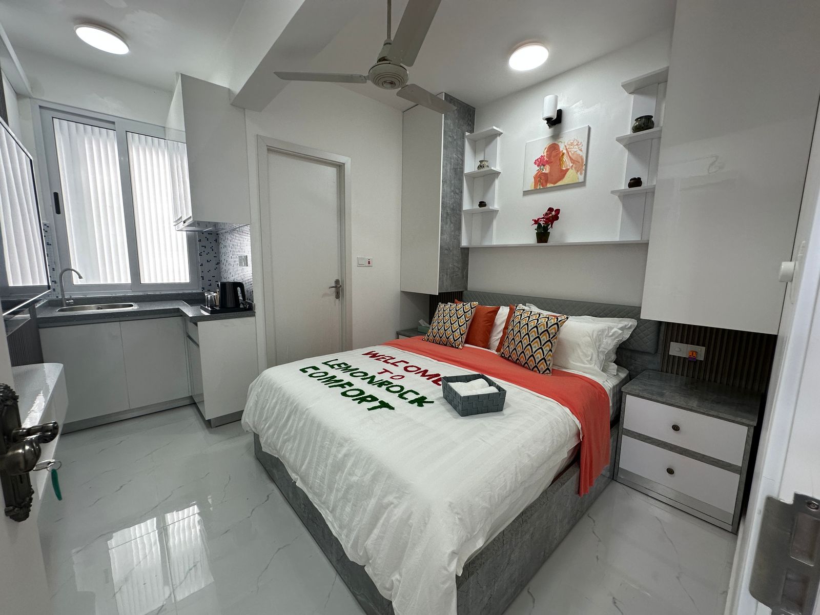 Modern bedroom with kitchenette, gray and white decor, and a bed with a welcome message on the duvet.