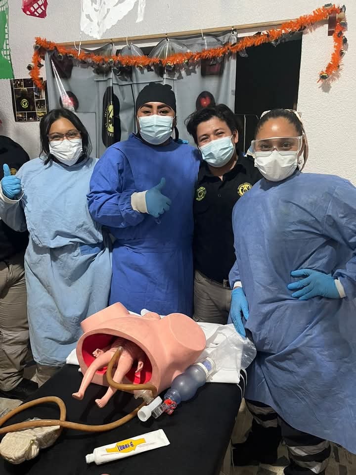Group of medical students in protective gear stand around a childbirth simulator model in a classroom.
