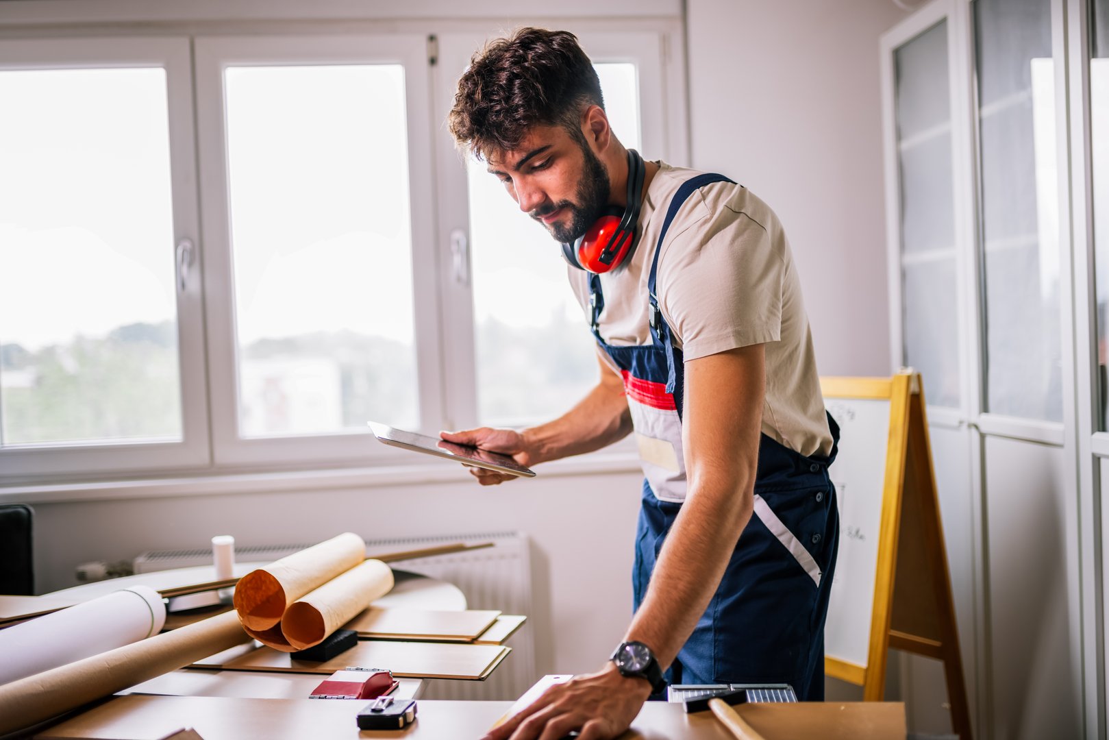 Repairman repairs apartment flooring, surrounded by laminate boards, and uses a tablet for guidance, combining modern technology with skilled craftsmanship.