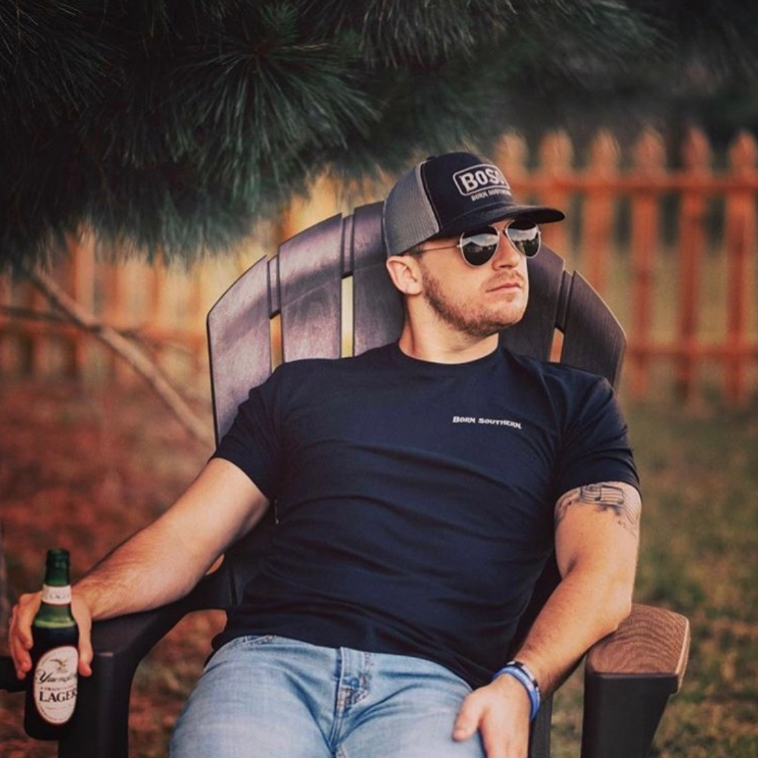 Man in sunglasses and cap relaxing in a chair outdoors, holding a beer bottle, with a wooden fence in the background.