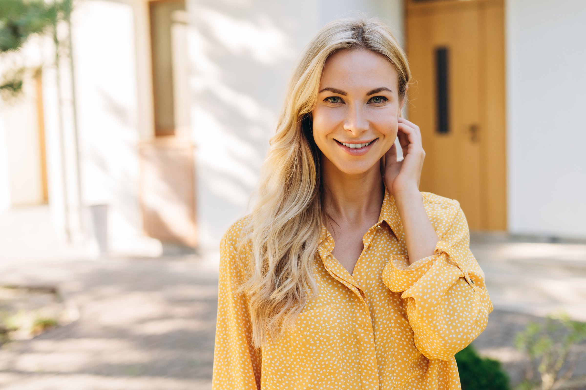 Portrait of a beautiful smiling blonde woman standing in front of her house, touching her hair and looking at camera