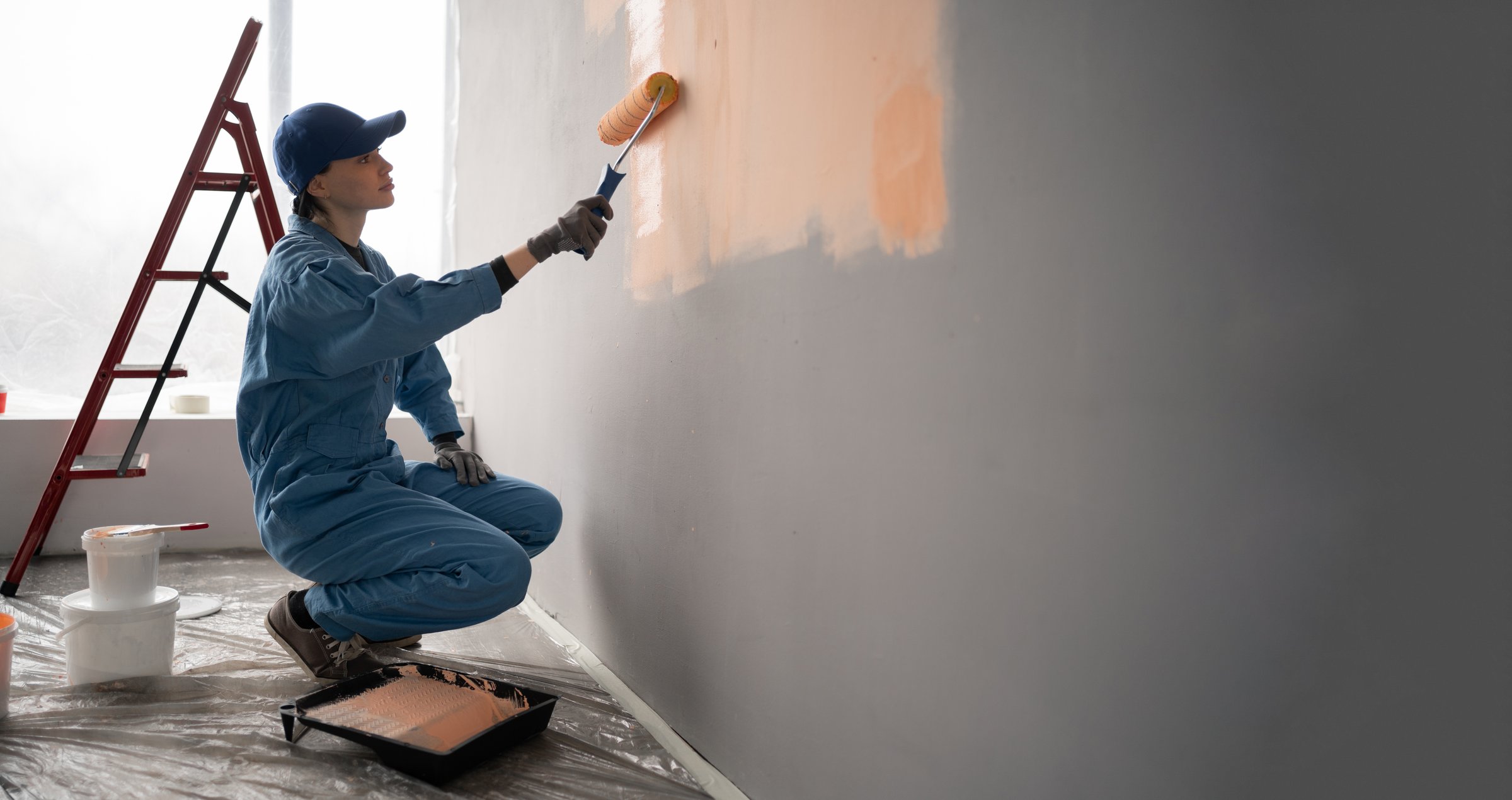 female painter in overalls with cap and gloves painting a wall with paint roller, ladder and buckets on background. Side view. Copy space