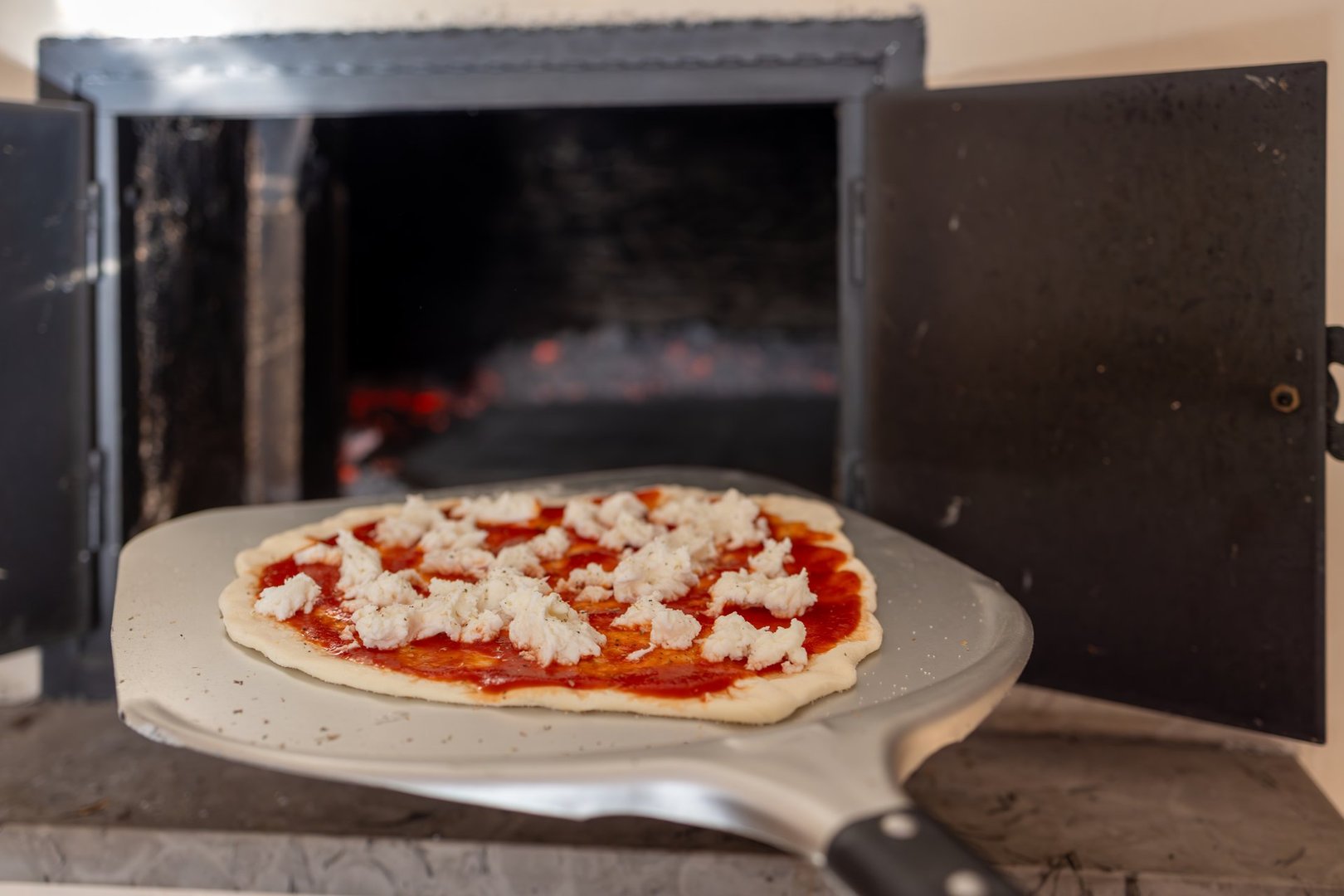A close-up of a freshly prepared pizza on a peel, ready to be placed inside a wood-fired oven with glowing embers. The image highlights the traditional pizza-making process and the inviting charm of artisanal cooking.