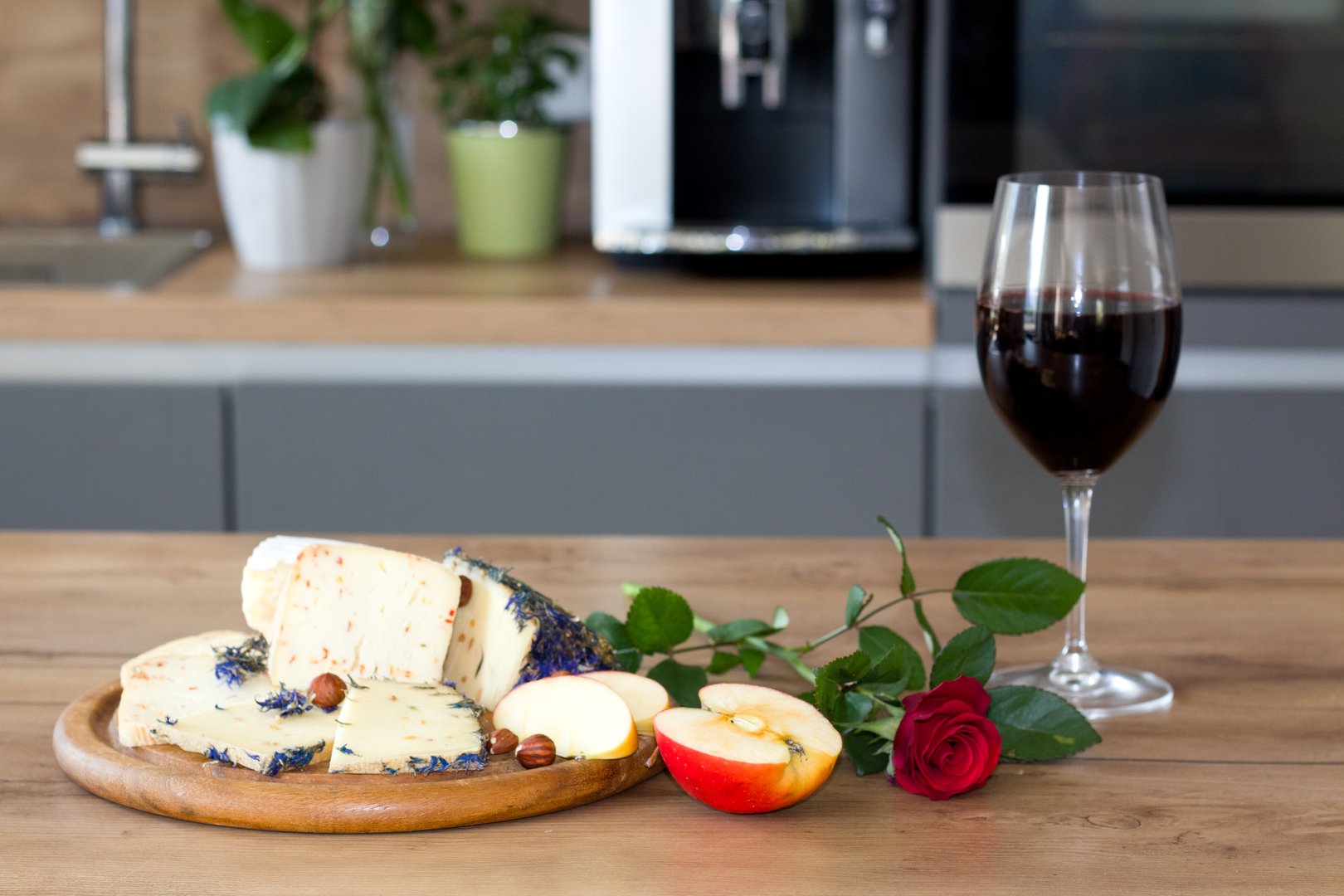 A delightful pairing of red wine, cheese adorned with edible flowers and nuts, apple slices, and a vibrant red rose on a rustic wooden board