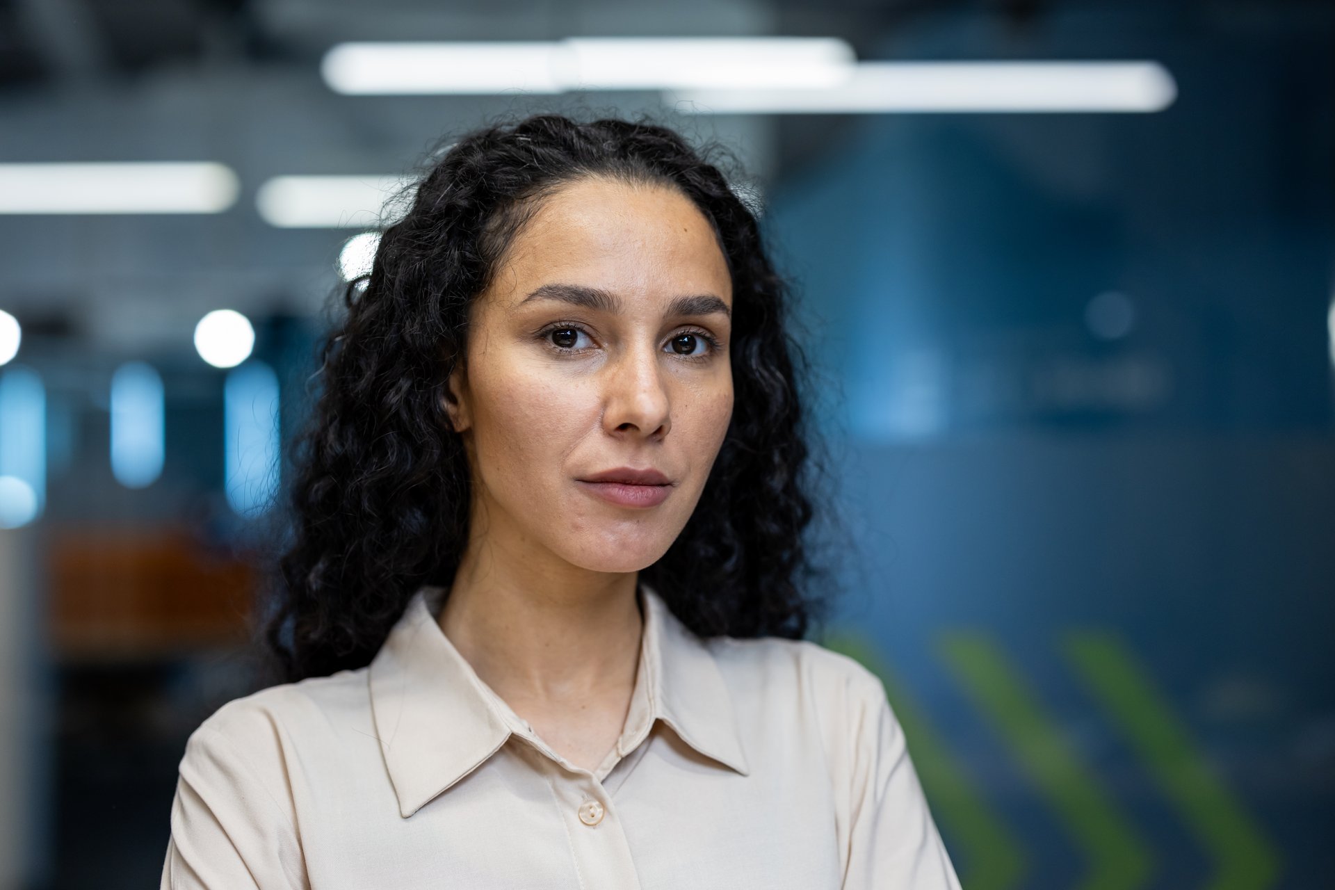 Portrait of confident businesswoman with curly hair standing in modern office. Professional woman wearing beige shirt, looking at camera with serious expression