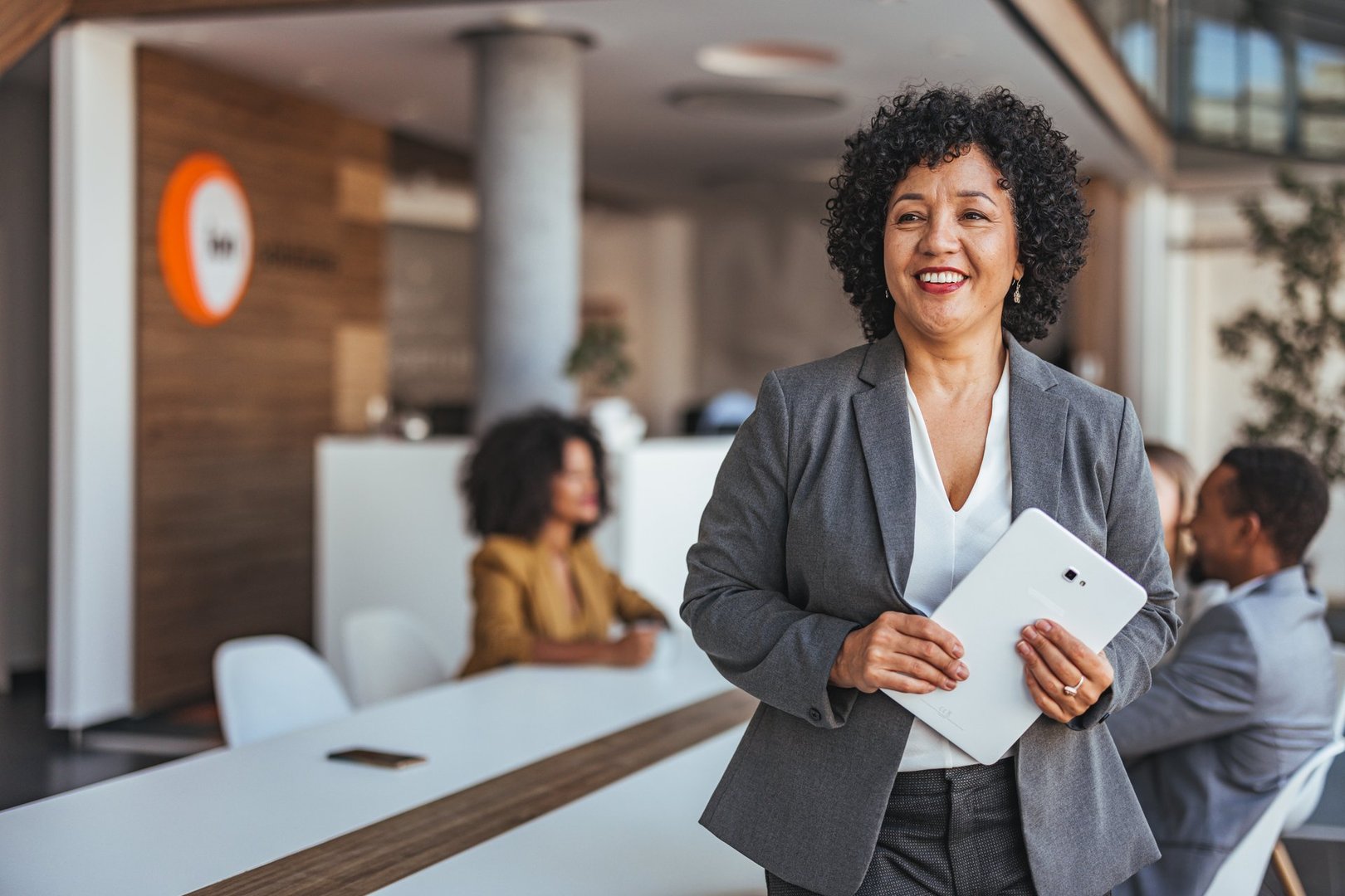 A confident businesswoman smiles while holding a tablet in a modern office environment. Colleagues are seen in the background, emphasizing teamwork and productivity.