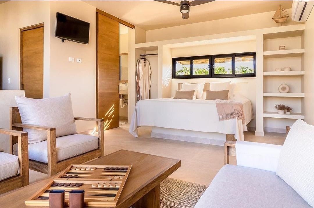 Modern bedroom with a cozy bed, armchairs, backgammon board, and wooden accents, featuring a sliding door and wall-mounted TV.