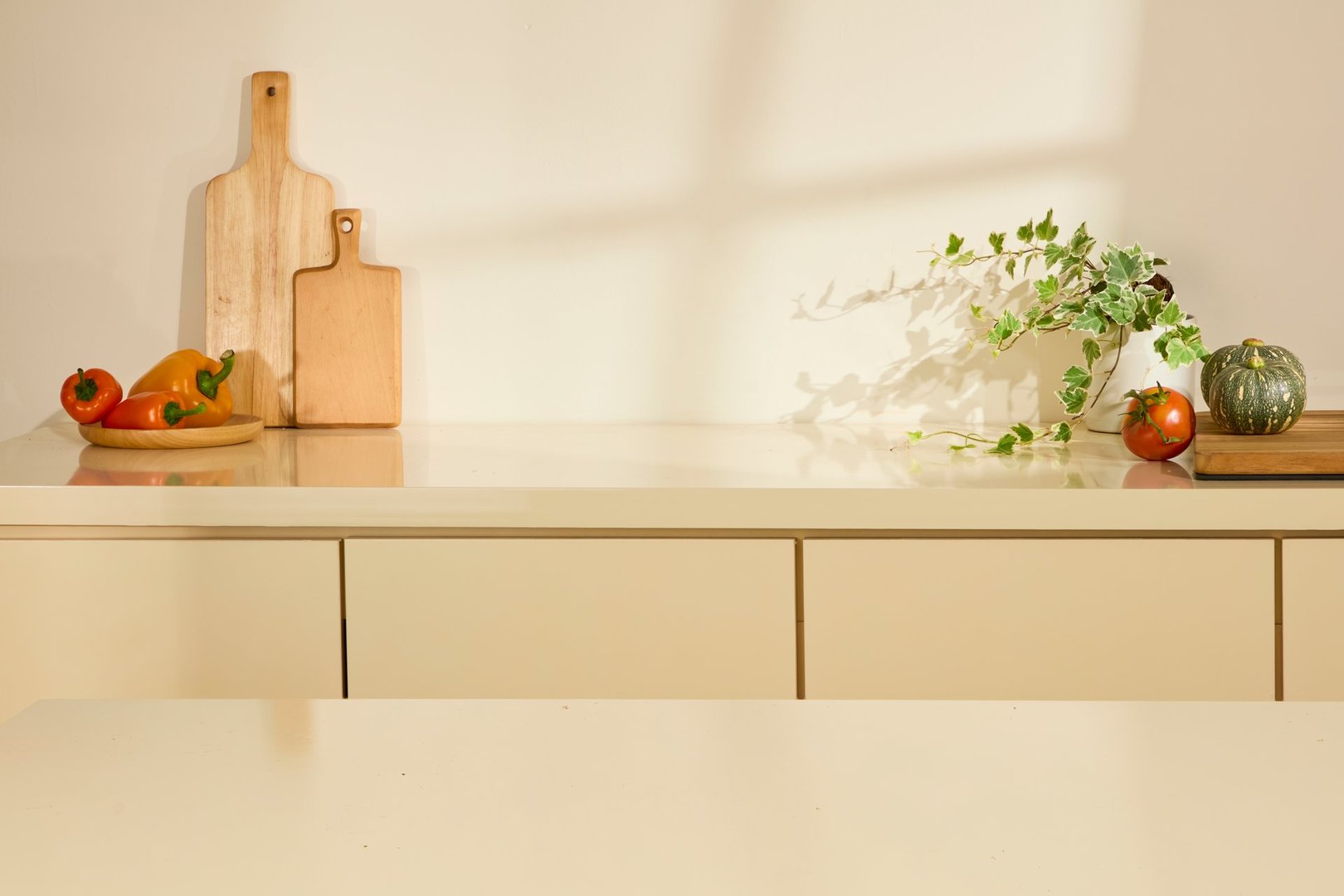 An empty dining table is used to place food or objects. Kitchen utensils are neatly arranged on the counter behind. Natural light shines through the window, leaving shadows on the wall. Copy space.