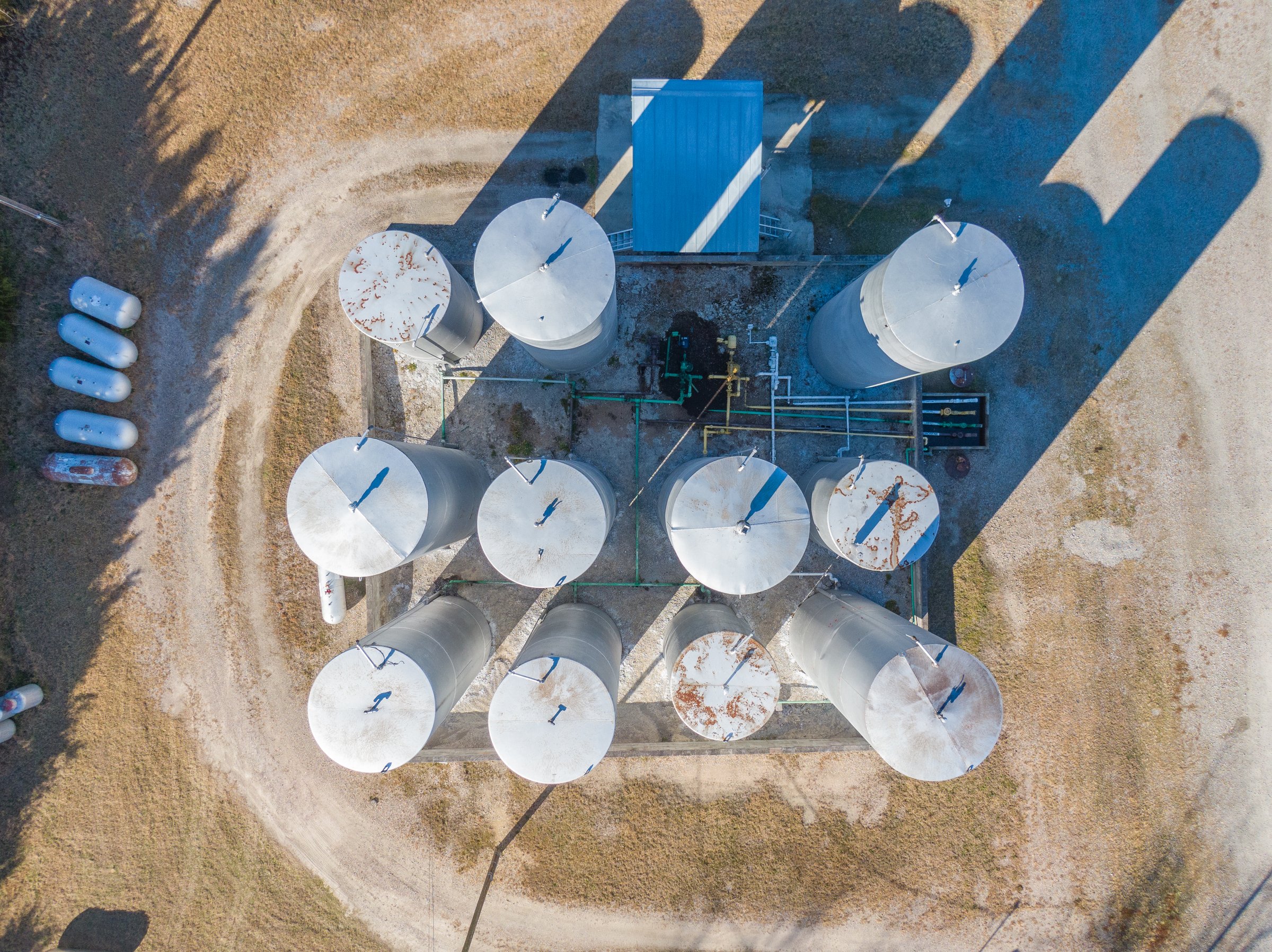 High-angle drone shot capturing a cluster of large vertical gasoline and diesel storage tanks with interconnected piping and weathered metal of the fuel facility.