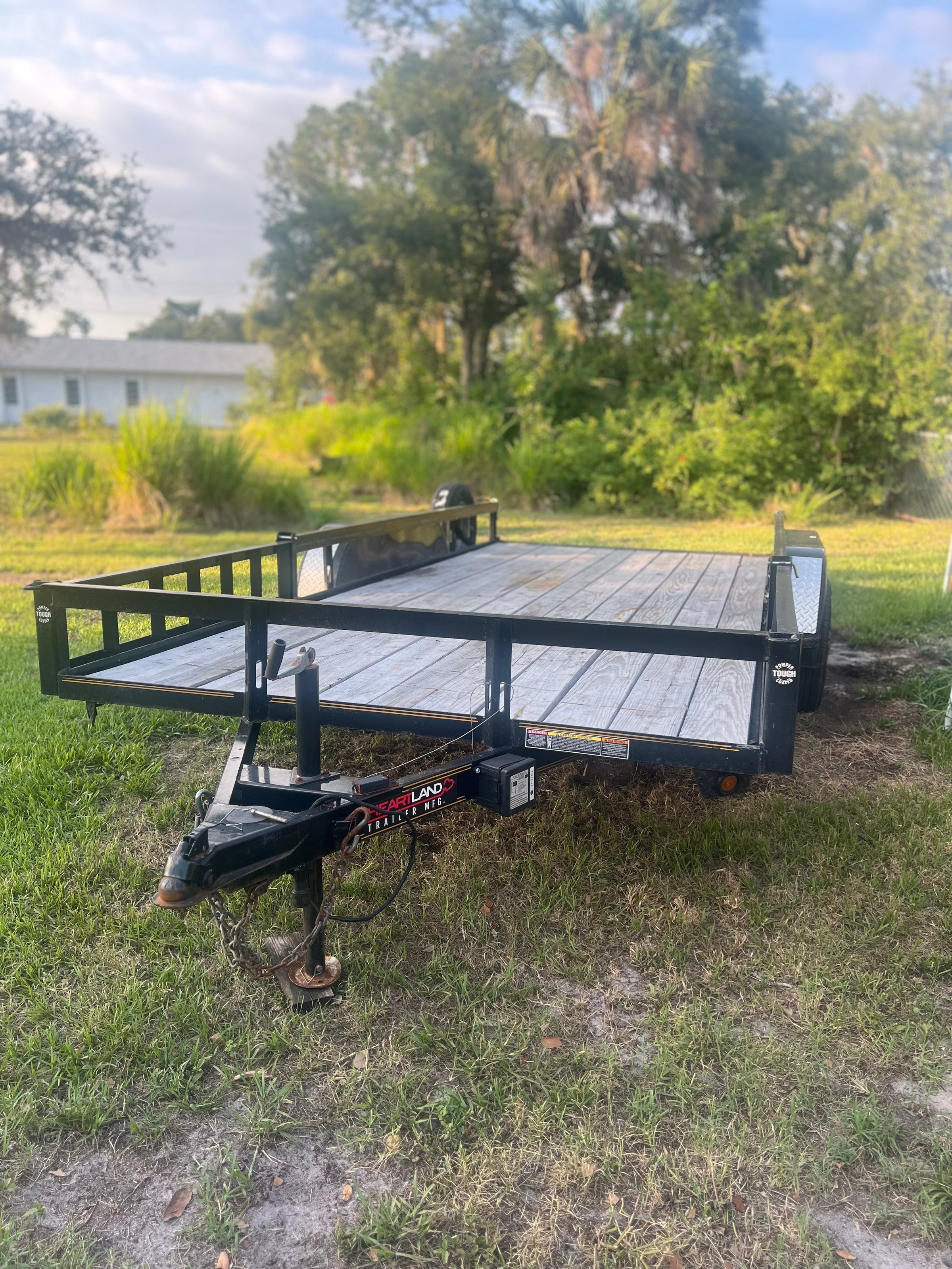 Utility trailer parked on grass with a wooden deck and metal frame, surrounded by trees and vegetation.