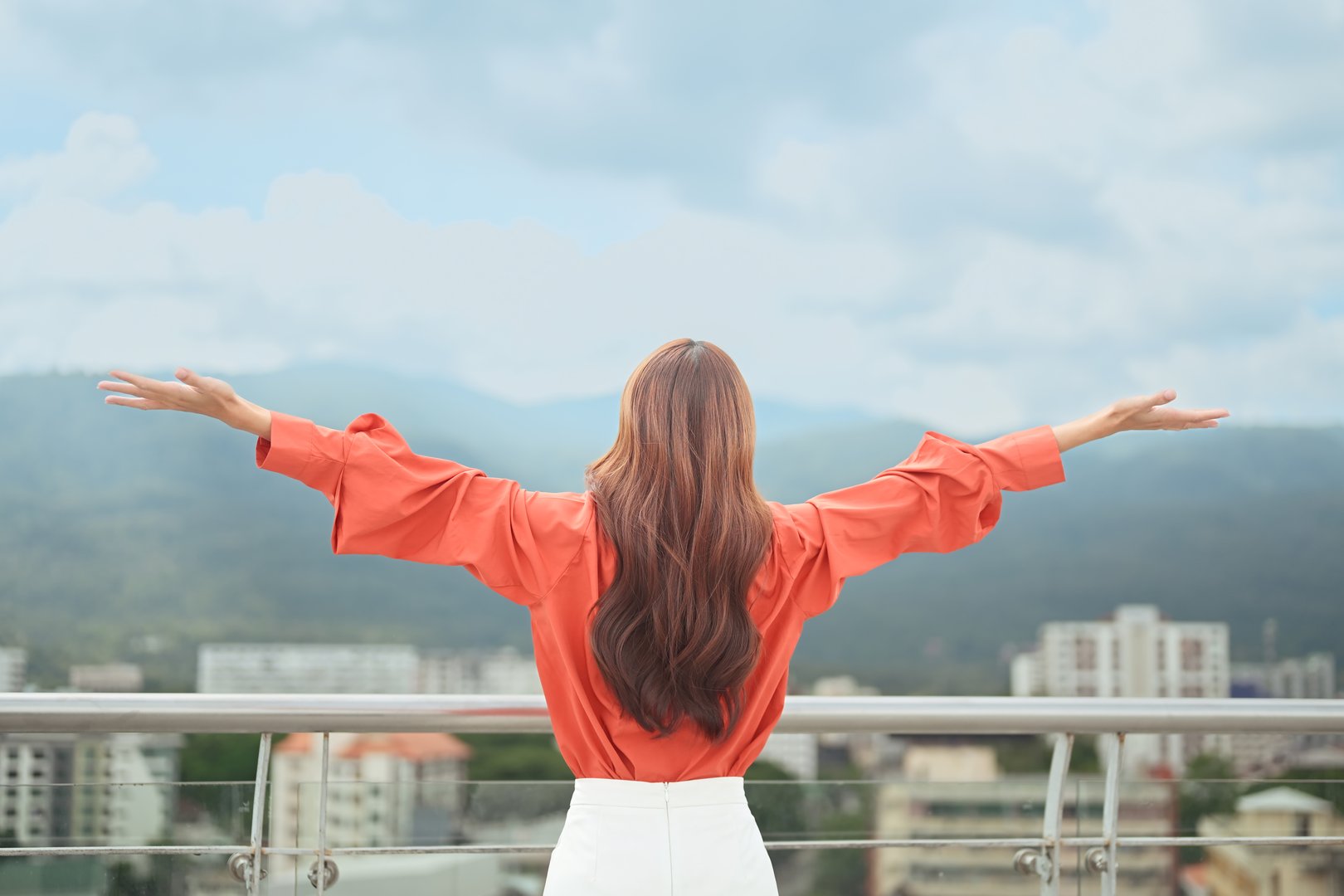 Back view of woman raising arms in freedom and happiness while standing on rooftop with city and mountain view.