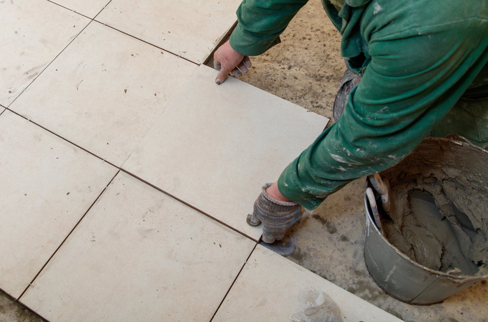A man is laying tiles on a floor. He is wearing a green shirt and gloves. The tiles are white and the floor is made of concrete