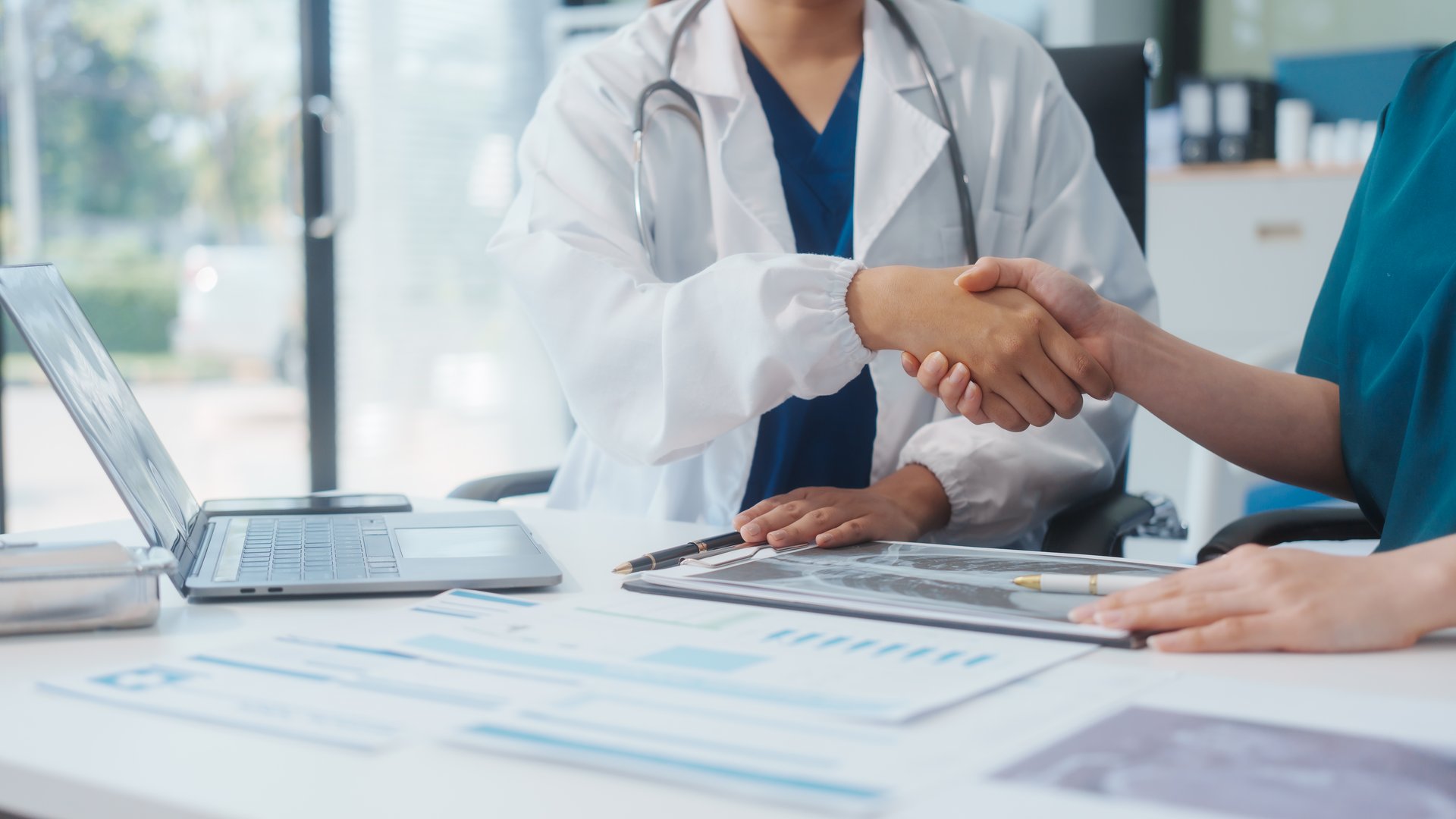 Two Asian female doctors shake hands after a successful meeting at a hospital, celebrating cooperation between two hospitals and recognizing their teamwork in advancing healthcare and patient care