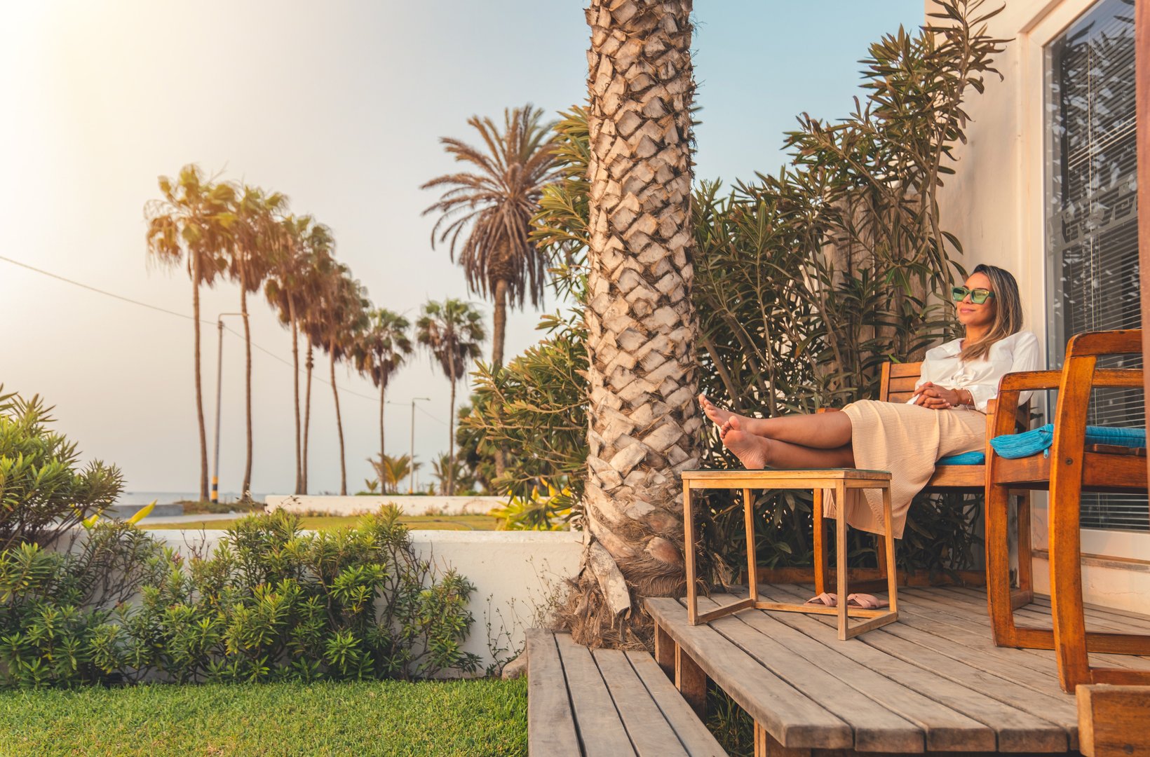A woman is sitting on a wooden bench outside of a house, with a palm tree in the background. She is wearing a white shirt and a pair of sunglasses. Concept of relaxation and leisure