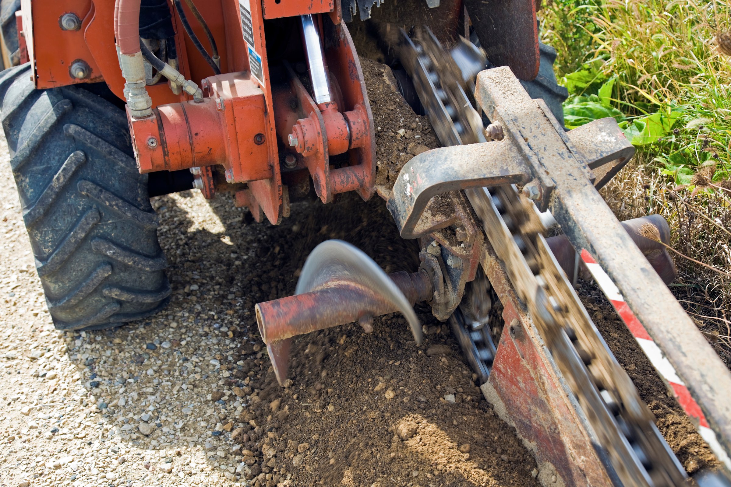 Trencher machine digging a trench in gravel, with rotating blade and moving chain visible.