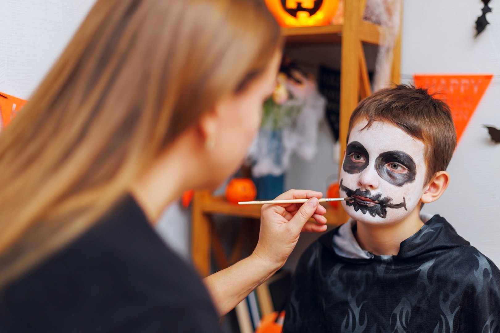 Woman applying skeleton face paint on a boy&#039;s face for Halloween celebration. The boy is dressed in a dark costume with Halloween decorations in the background