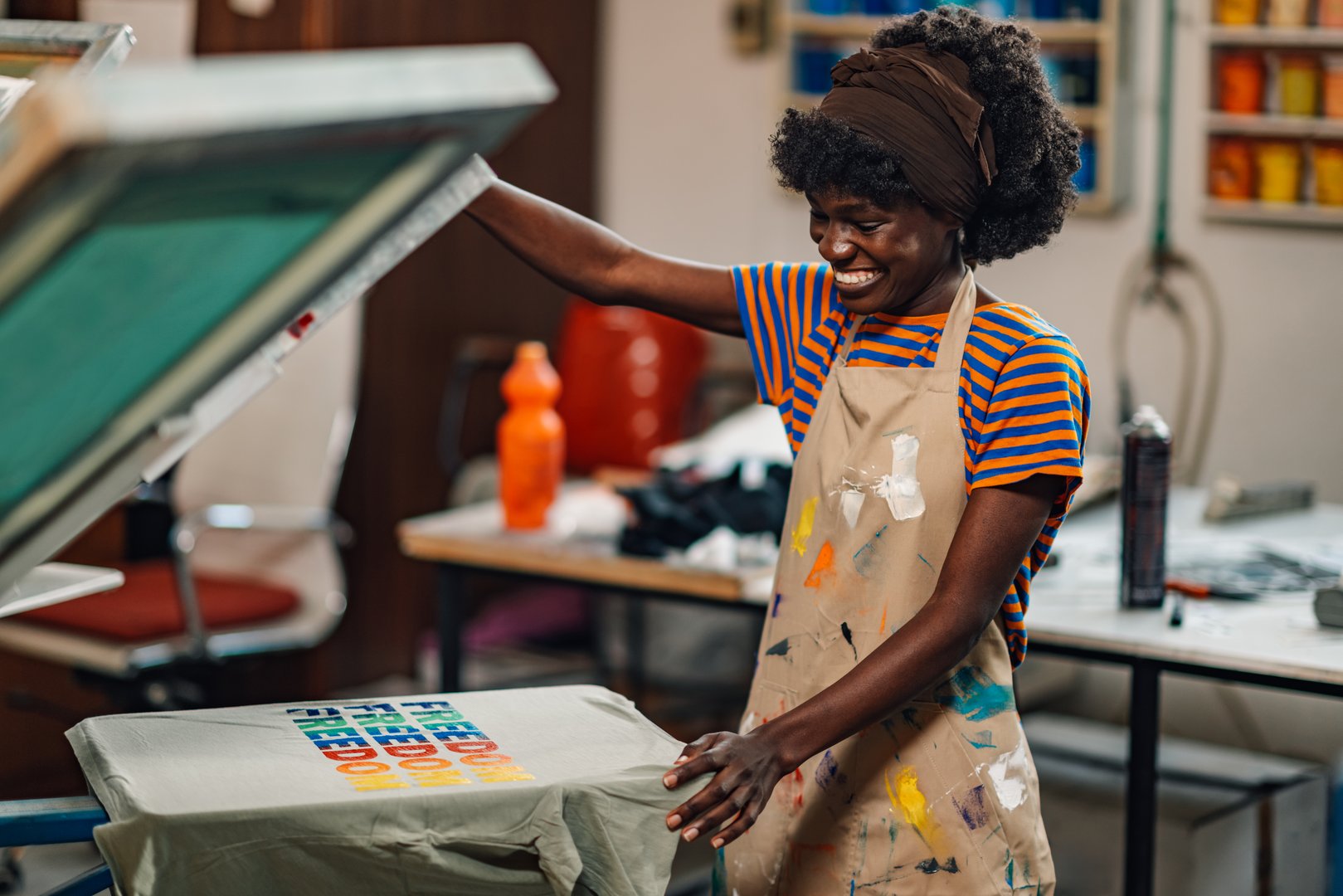 Happy african american professional graphic technician is finishing silk screen printing on t-shirt at printing shop. Smiling multicultural graphic technology expert is smiling at finished product.