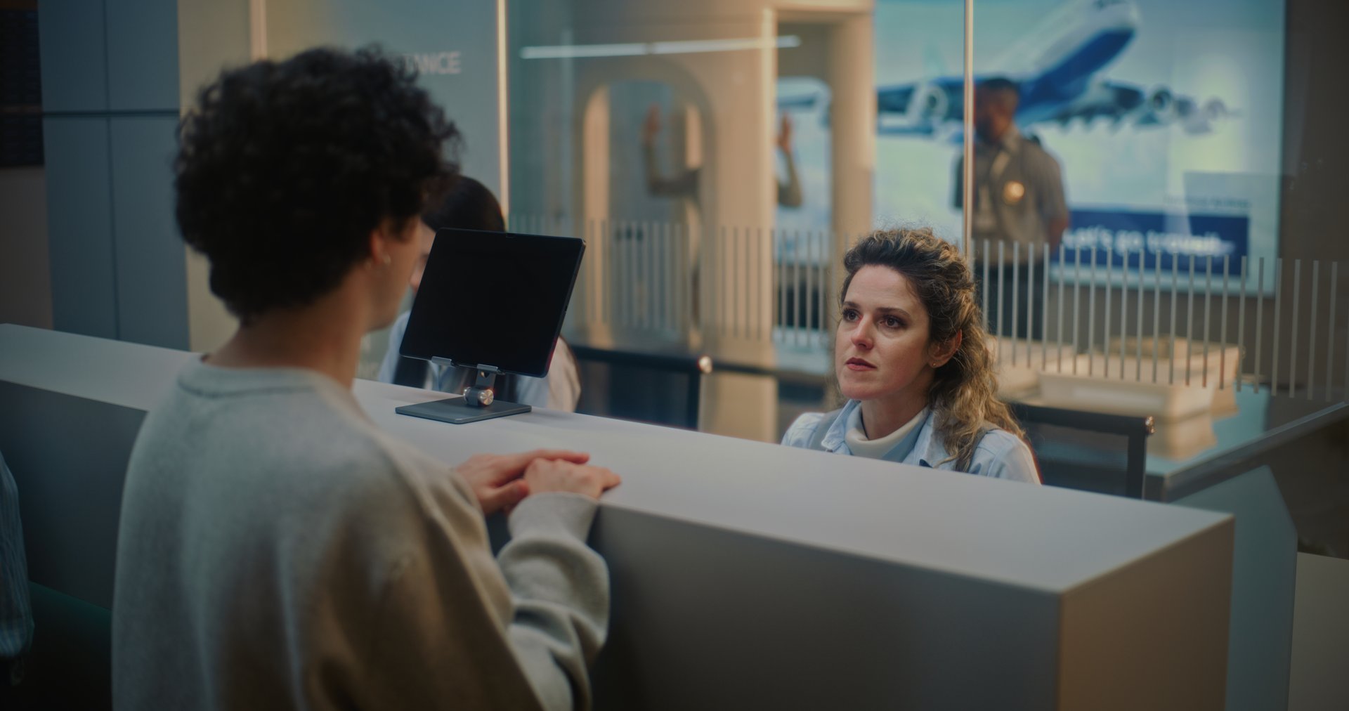 Airport Check-in Counter in International Airport Terminal: Female Airline Worker Talking to Young Man, Asking Questions During Passport Check for Boarding Flight. Security Checkpoint in Background.