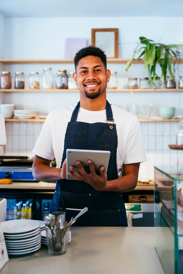 Bakery, happy portrait of hispanic black man in cafe ready for serving pastry, coffee and baked foods. Restaurant, coffee shop and confident waiter barista by counter for service, help and welcome