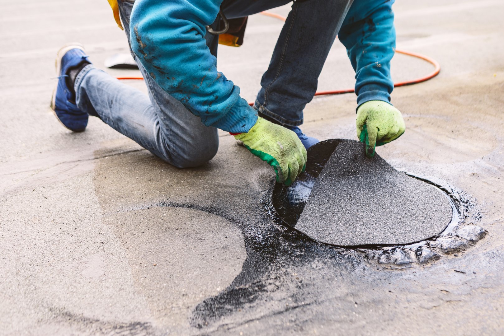 Roofer repairing a damaged section of a roof. Worker kneels on a wet, flat rooftop to apply a circular patch. Their gloved hands press the black material into place, sealing a roof