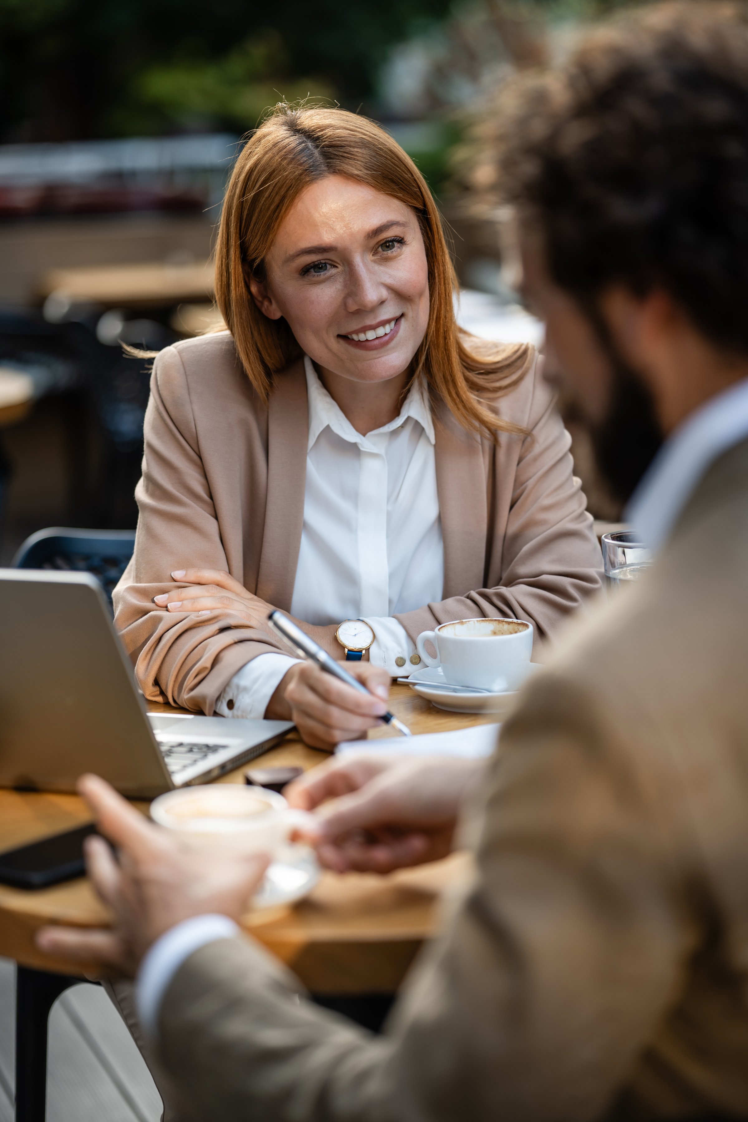 Businesswoman smiling and taking notes during a meeting with a male colleague at an outdoor cafe, enjoying coffee and discussing work