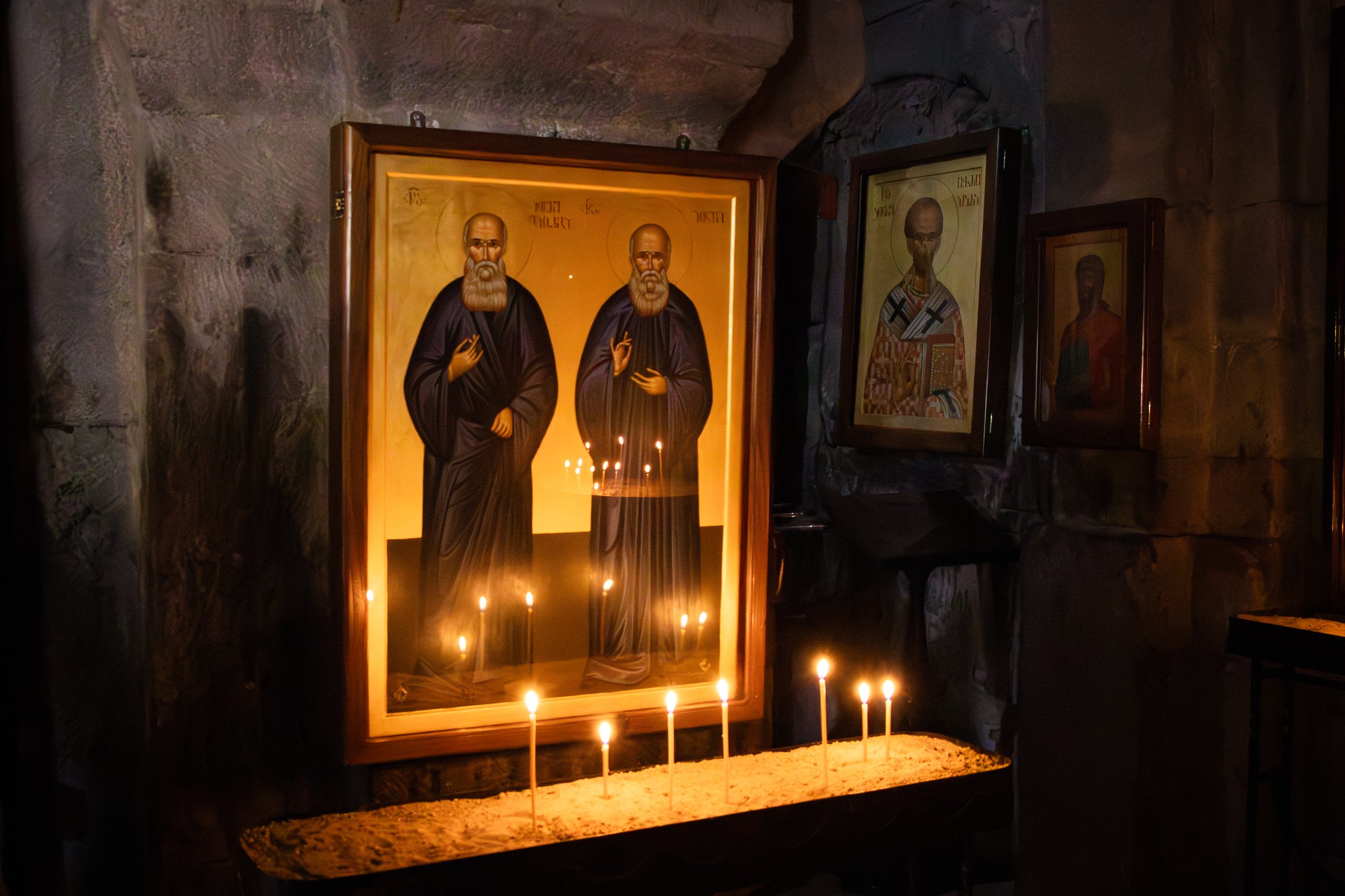 Sacred atmosphere inside the Gergeti Trinity Monastery Church in Georgia. Icon paintings of saints are illuminated by the soft glow of candlelight, casting warm reflections on the ancient stone walls. A serene representation of faith, prayer, and tradition in this historic Caucasus landmark.