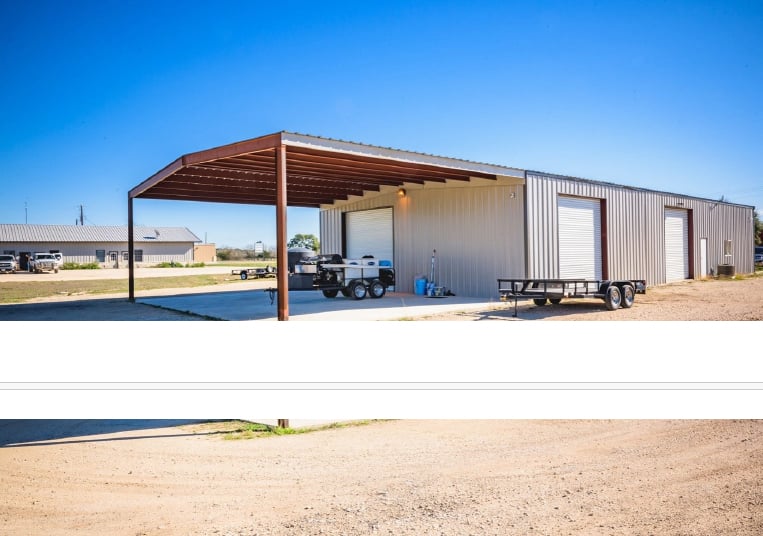 Large metal building with open-sided shelter, trailers parked underneath, and clear blue sky background.