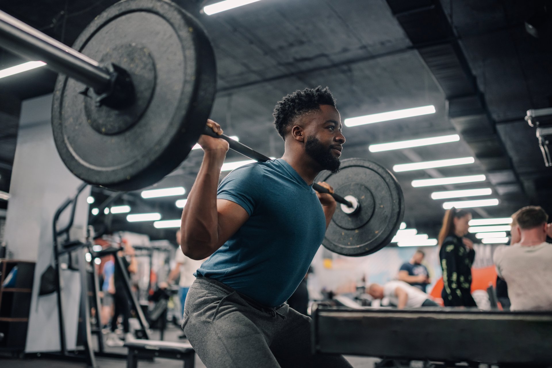 Young muscular athlete doing squats with barbell in a gym, surrounded by other people training