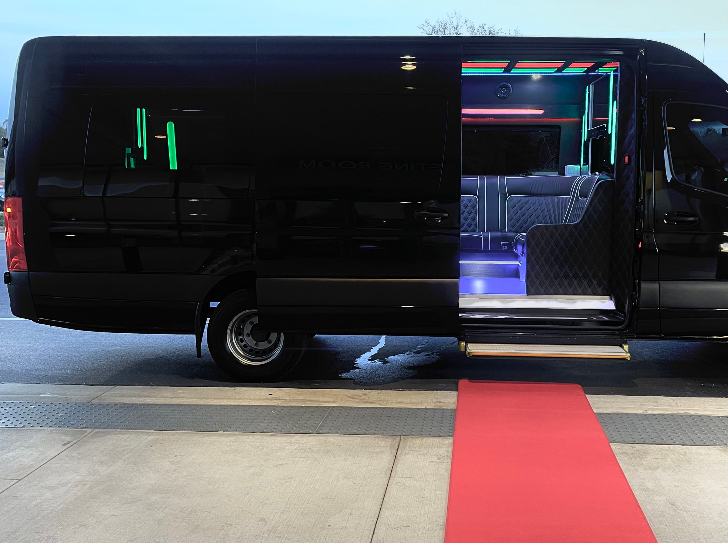 Black luxury van with open door, revealing neon-lit interior. A red carpet extends to the entrance.
